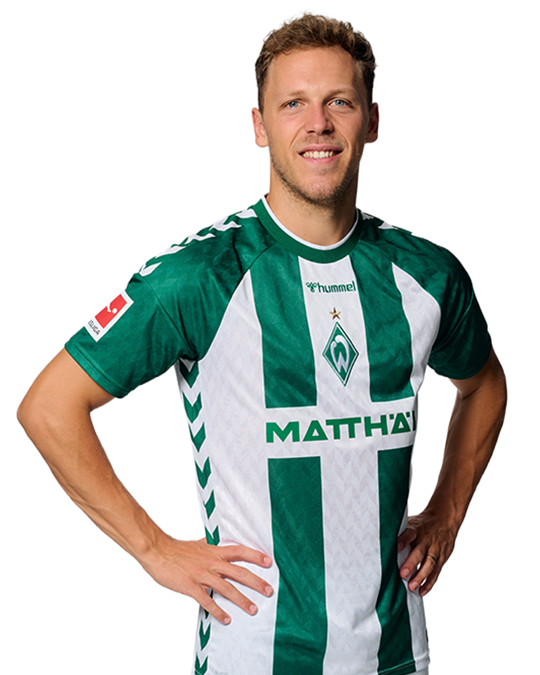 Ein Mann in einem grün-weiß gestreiften Fußballtrikot von Werder Bremen steht selbstbewusst mit den Händen in den Hüften vor einem einfarbigen Hintergrund.