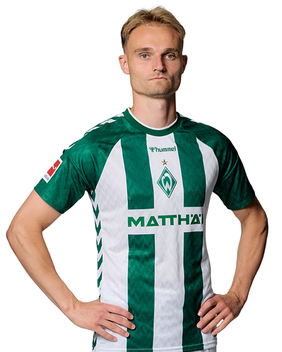 Ein Mann in einem grün-weiß gestreiften Fußballtrikot von Werder Bremen steht selbstbewusst mit den Händen in den Hüften vor einem einfarbigen Hintergrund.
