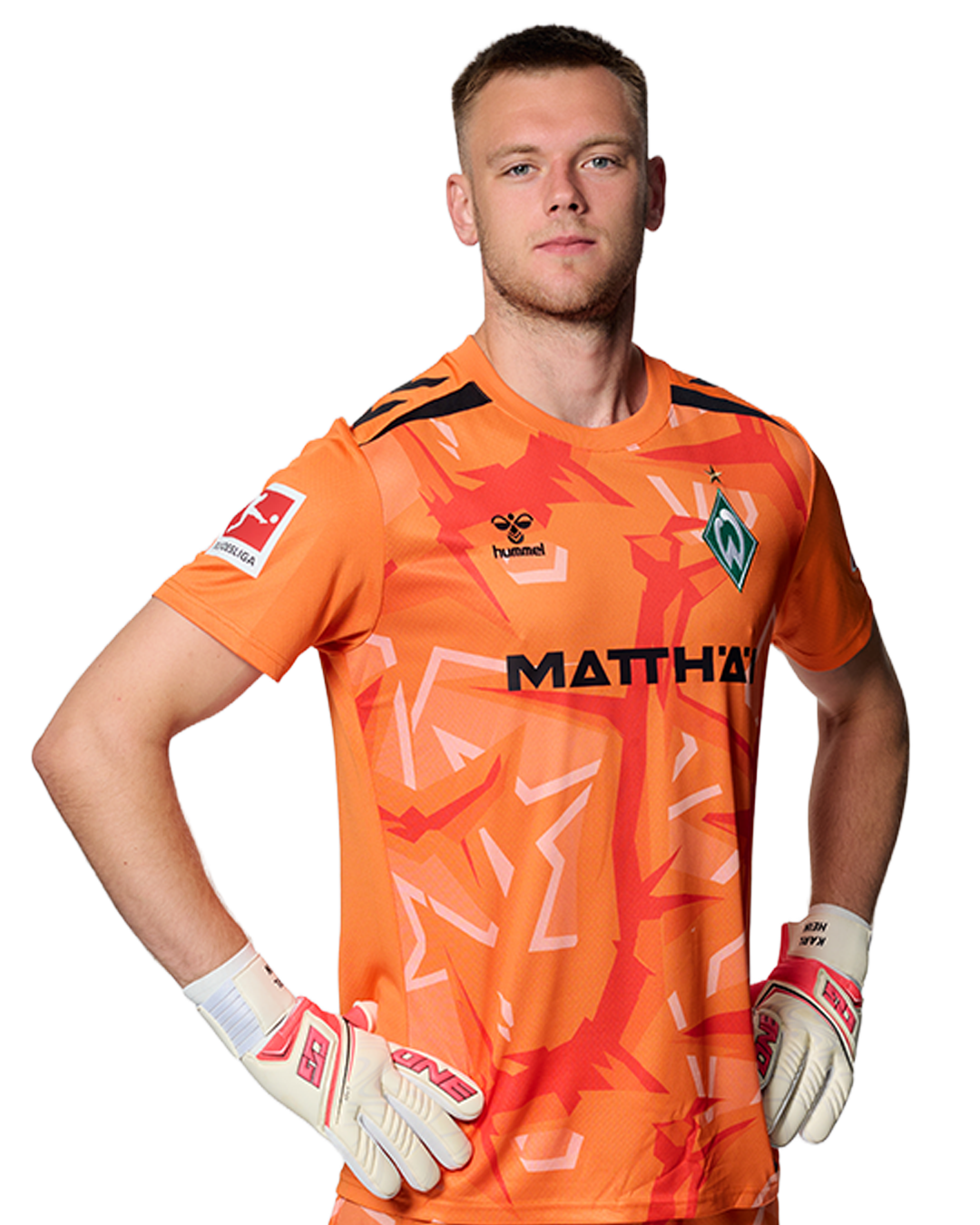 Ein Mann in einem orangenen gestreiften Fußballtrikot von Werder Bremen steht selbstbewusst mit den Händen in den Hüften vor einem einfarbigen Hintergrund.