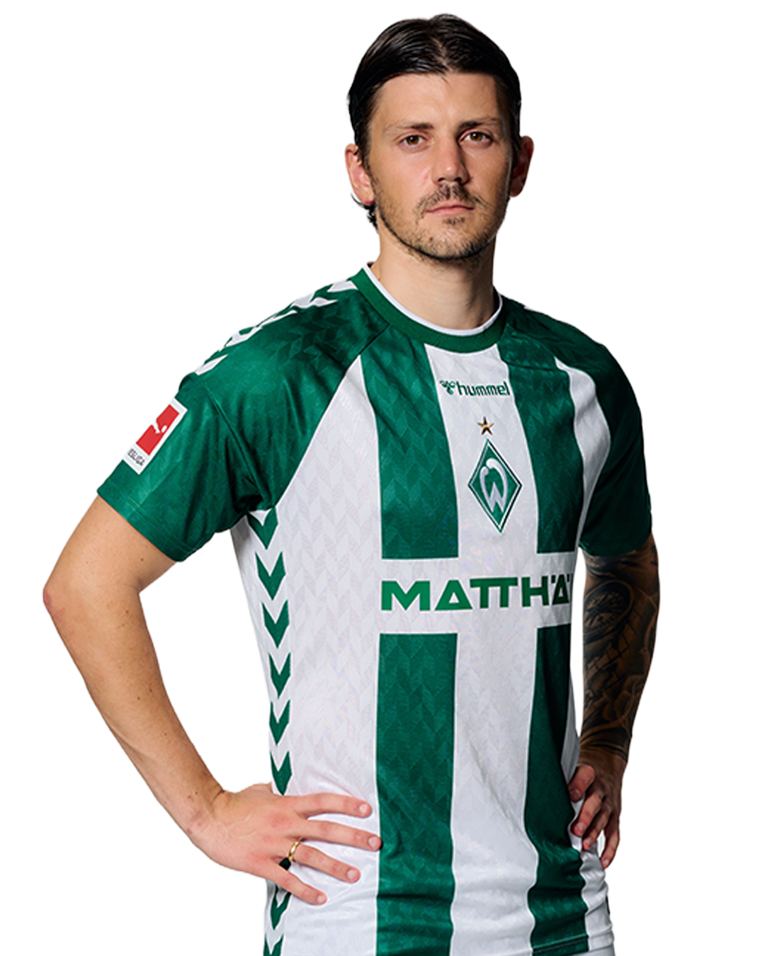 Ein Mann in einem grün-weiß gestreiften Fußballtrikot von Werder Bremen steht selbstbewusst mit den Händen in den Hüften vor einem einfarbigen Hintergrund.