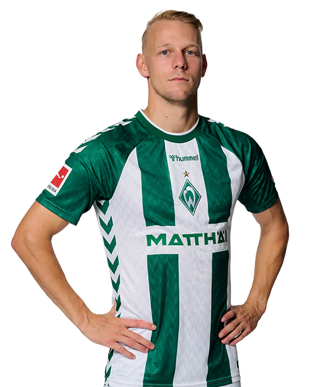 Ein Mann in einem grün-weiß gestreiften Fußballtrikot von Werder Bremen steht selbstbewusst mit den Händen in den Hüften vor einem einfarbigen Hintergrund.