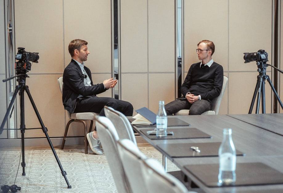 Zwei Männer sitzen in einem Konferenzraum und unterhalten sich, während sie von professionellen Kameras gefilmt werden. Auf dem Tisch stehen eine Wasserflasche und Notizbücher.