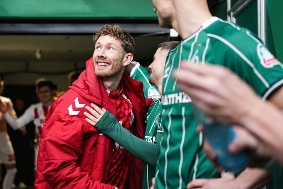 Fußballspieler in grünen und roten Trikots lächeln und machen sich bereit, aus dem Spielertunnel auf den Platz zu laufen, wodurch eine Atmosphäre der Vorfreude und Kameradschaft entsteht.