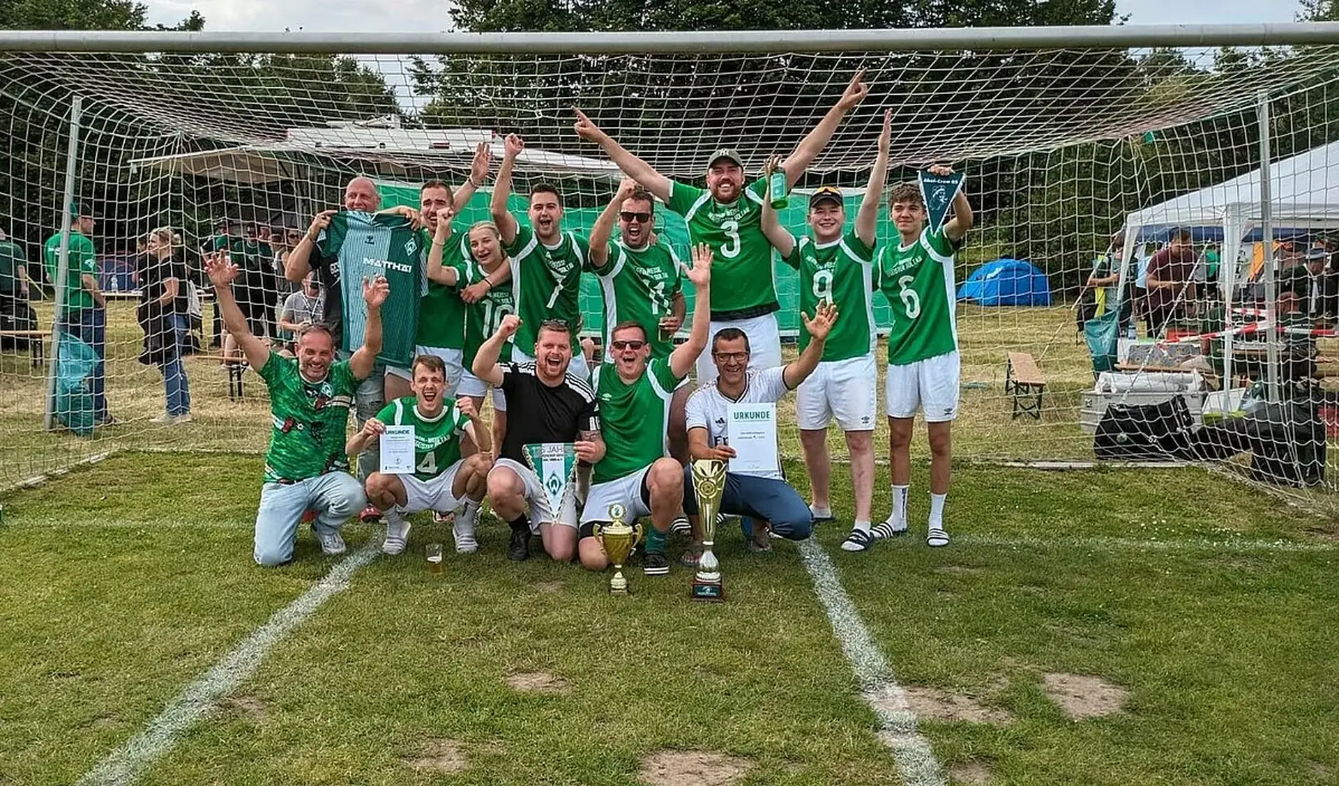 Werder-Fans jubeln bei der Fanclub-Meisterschaft auf einem Gruppenfoto. Sie stehen auf einem Rasenplatz im Tor, vor ihnen stehen zwei Pokale.
