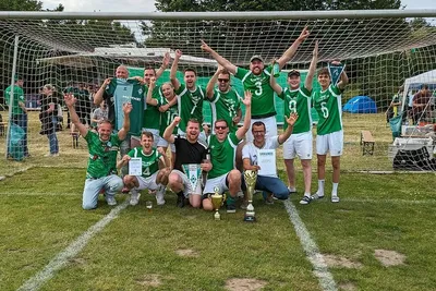 Werder-Fans jubeln bei der Fanclub-Meisterschaft auf einem Gruppenfoto. Sie stehen auf einem Rasenplatz im Tor, vor ihnen stehen zwei Pokale.