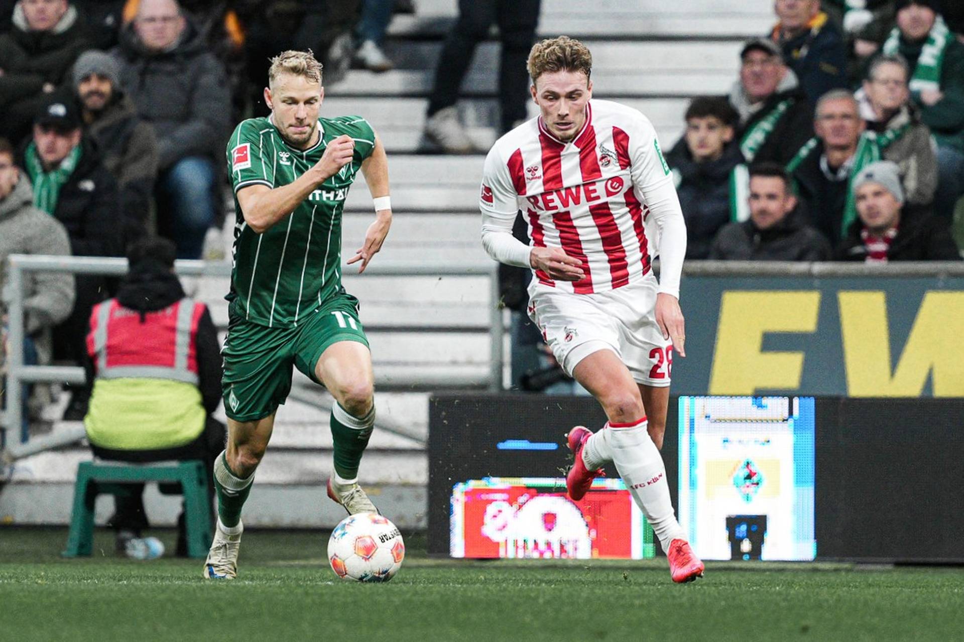 Zwei Fußballspieler in Bewegung auf dem Spielfeld; einer in grün gestreiftem Trikot, der andere in rot-weiß gestreiftem Trikot, mit Zuschauer*innen im Hintergrund.