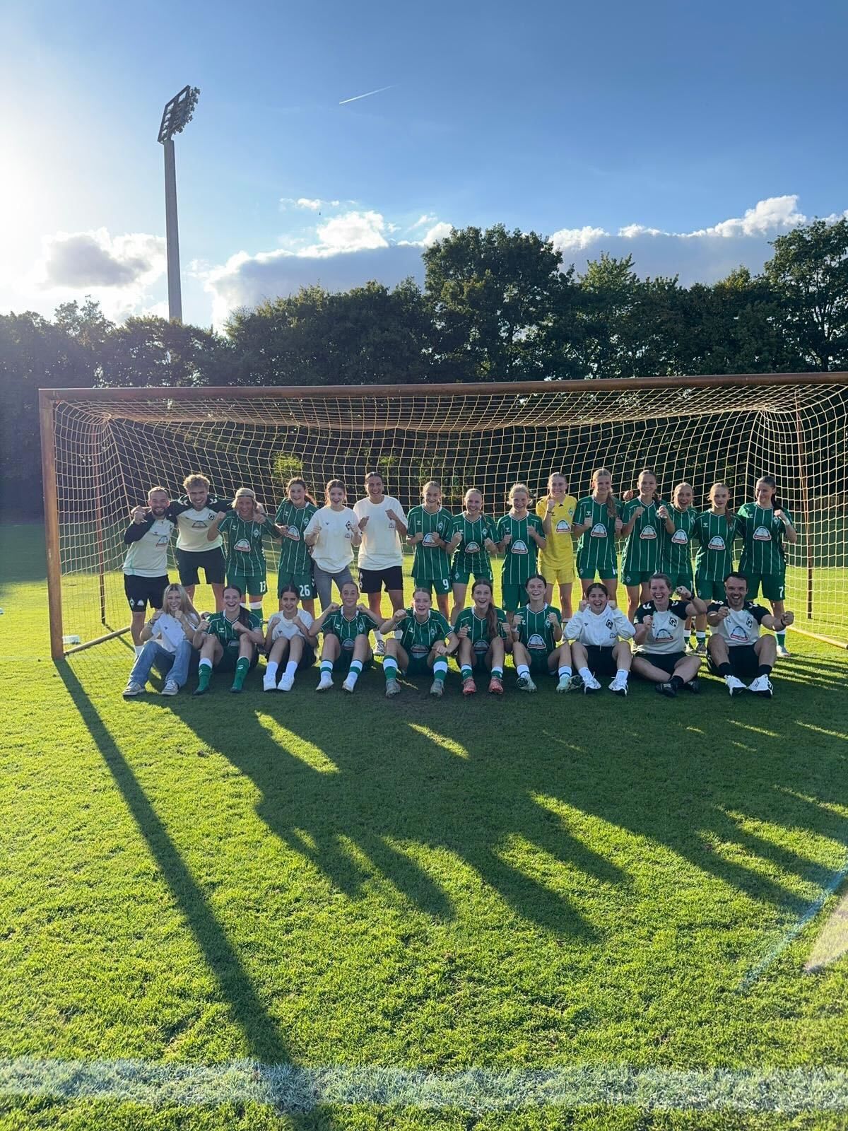 Die U17-Juniorinnen posieren in grünen Trikots gemeinsam auf einem Feld vor einem Tor, während die Sonne lange Schatten wirft.