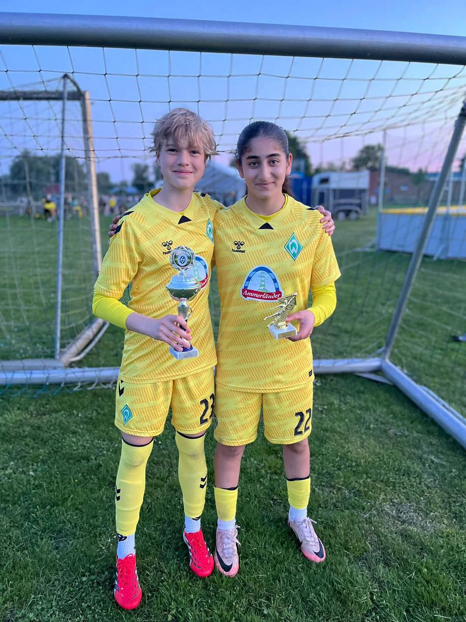 Zwei junge Fußballspielerinnen in gelben Trikots stehen auf einem Spielfeld, jede hält einen Pokal in der Hand und lächelt in die Kamera, hinter ihnen ist ein Netz zu sehen.