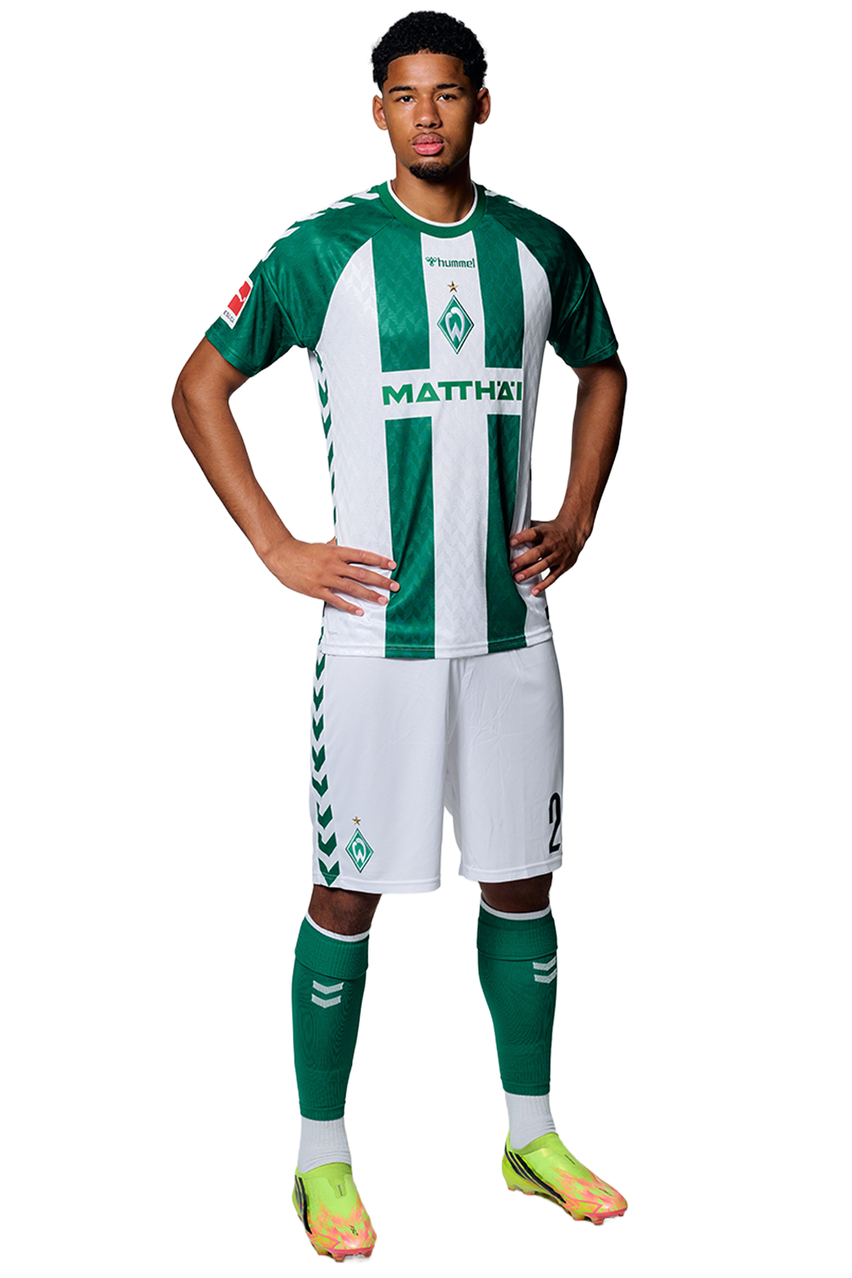 Ein Mann in einem grün-weiß gestreiften Fußballtrikot von Werder Bremen steht selbstbewusst mit den Händen in den Hüften vor einem einfarbigen Hintergrund.