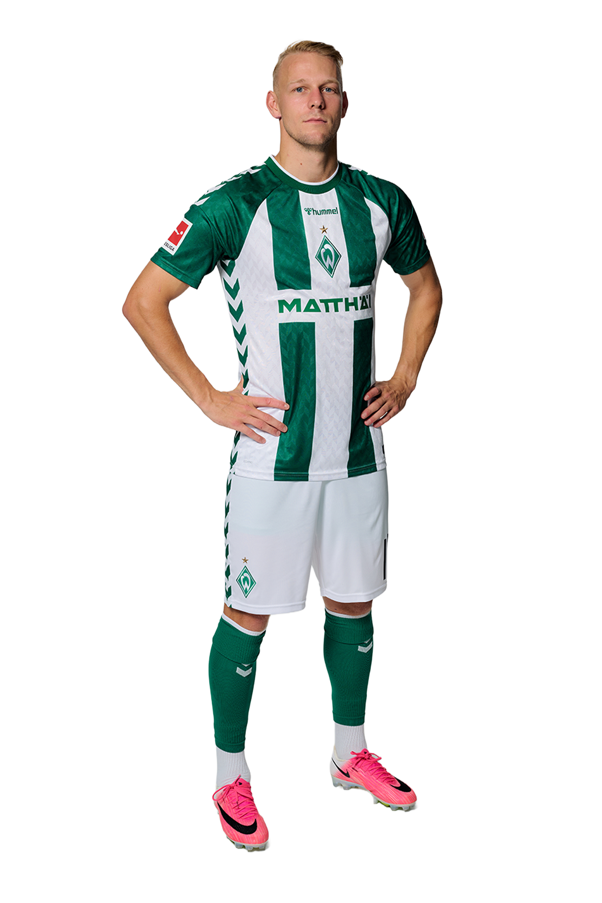Ein Mann in einem grün-weiß gestreiften Fußballtrikot von Werder Bremen steht selbstbewusst mit den Händen in den Hüften vor einem einfarbigen Hintergrund.