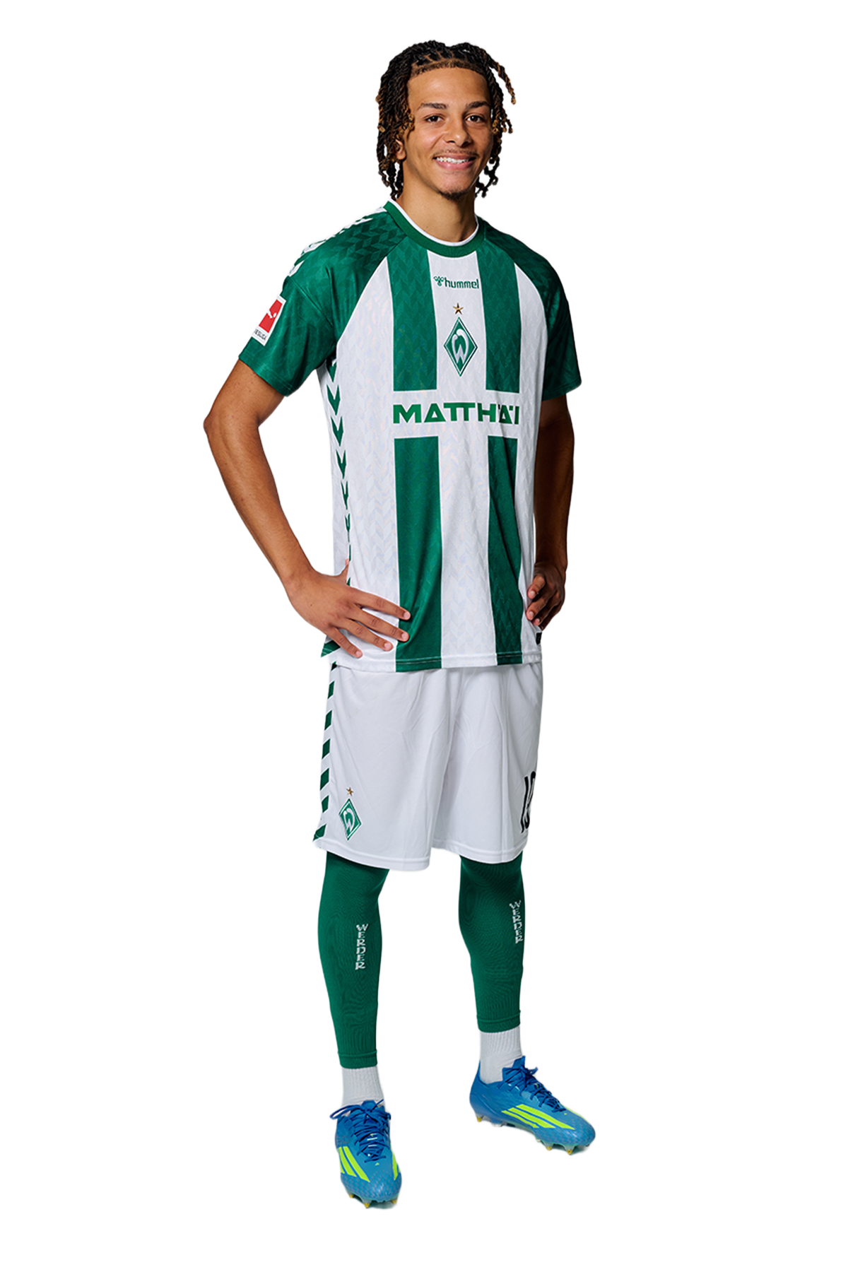 Ein Mann in einem grün-weiß gestreiften Fußballtrikot von Werder Bremen steht selbstbewusst mit den Händen in den Hüften vor einem einfarbigen Hintergrund.
