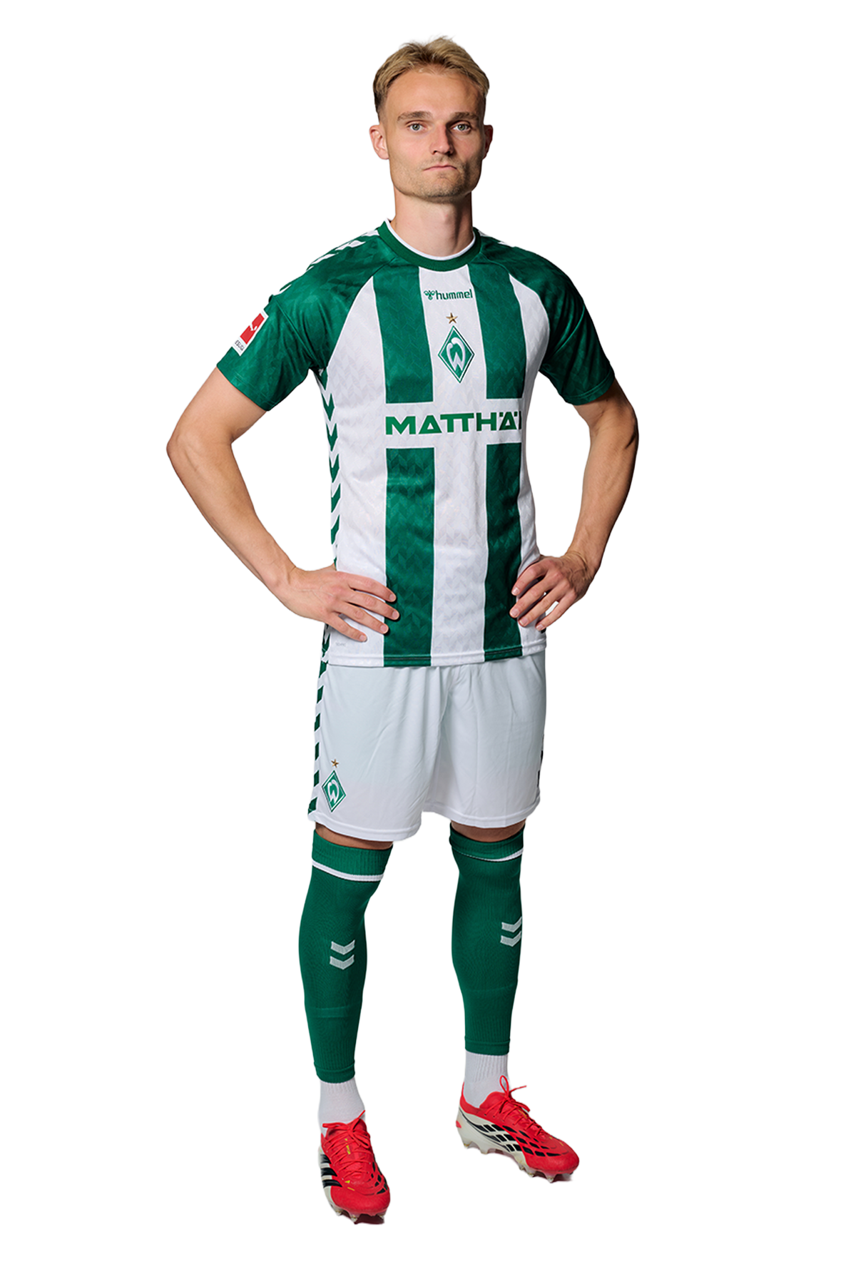 Ein Mann in einem grün-weiß gestreiften Fußballtrikot von Werder Bremen steht selbstbewusst mit den Händen in den Hüften vor einem einfarbigen Hintergrund.