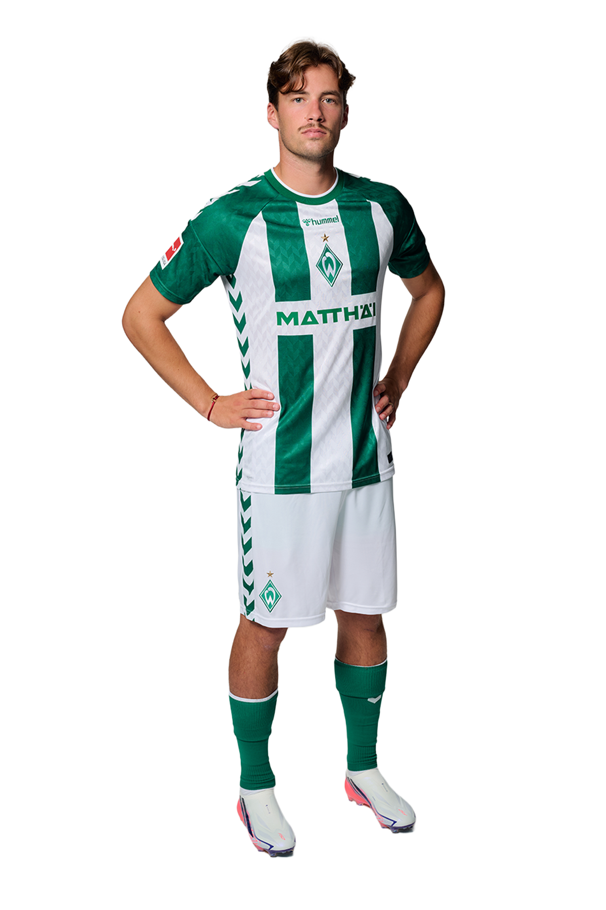 Ein Mann in einem grün-weiß gestreiften Fußballtrikot von Werder Bremen steht selbstbewusst mit den Händen in den Hüften vor einem einfarbigen Hintergrund.
