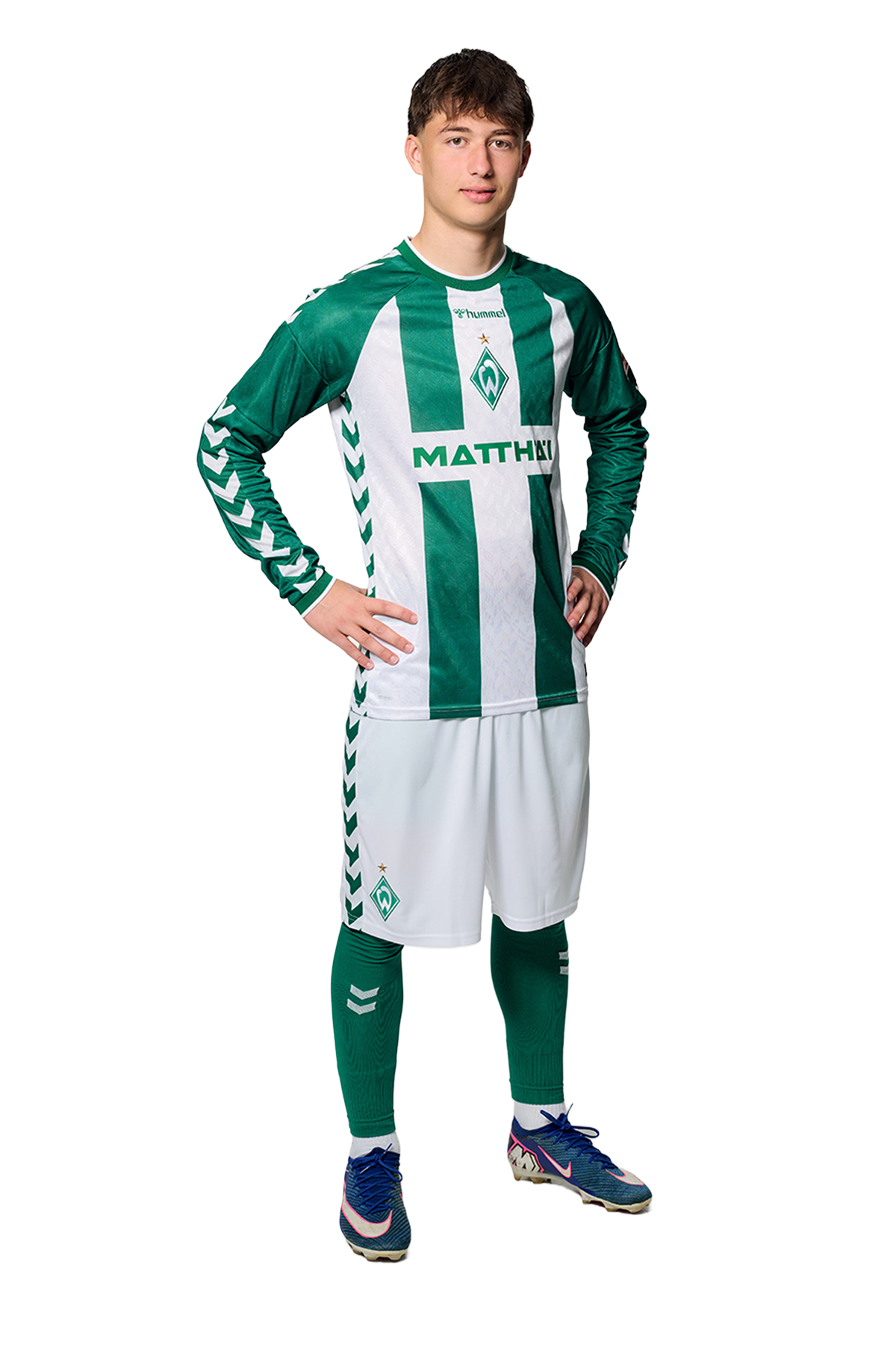 Ein Mann in einem grün-weiß gestreiften Fußballtrikot von Werder Bremen steht selbstbewusst mit den Händen in den Hüften vor einem einfarbigen Hintergrund.