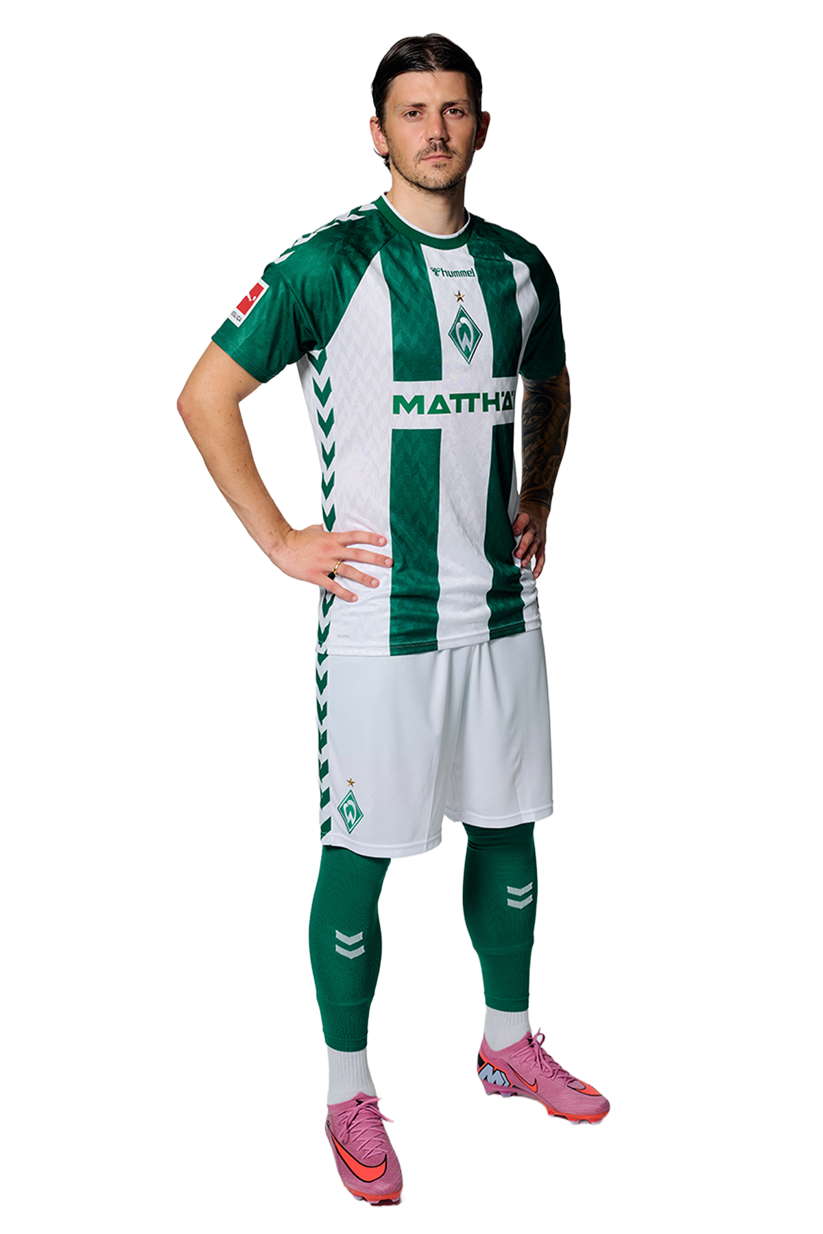 Ein Mann in einem grün-weiß gestreiften Fußballtrikot von Werder Bremen steht selbstbewusst mit den Händen in den Hüften vor einem einfarbigen Hintergrund.