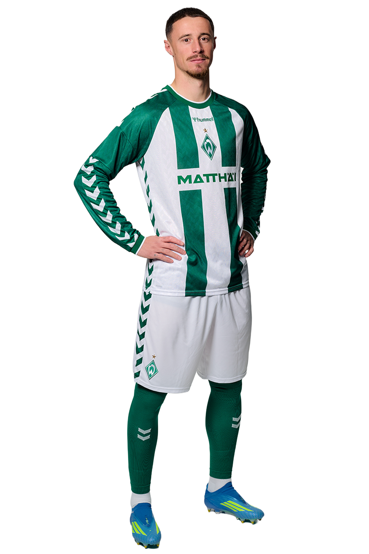 Ein Mann in einem grün-weiß gestreiften Fußballtrikot von Werder Bremen steht selbstbewusst mit den Händen in den Hüften vor einem einfarbigen Hintergrund.