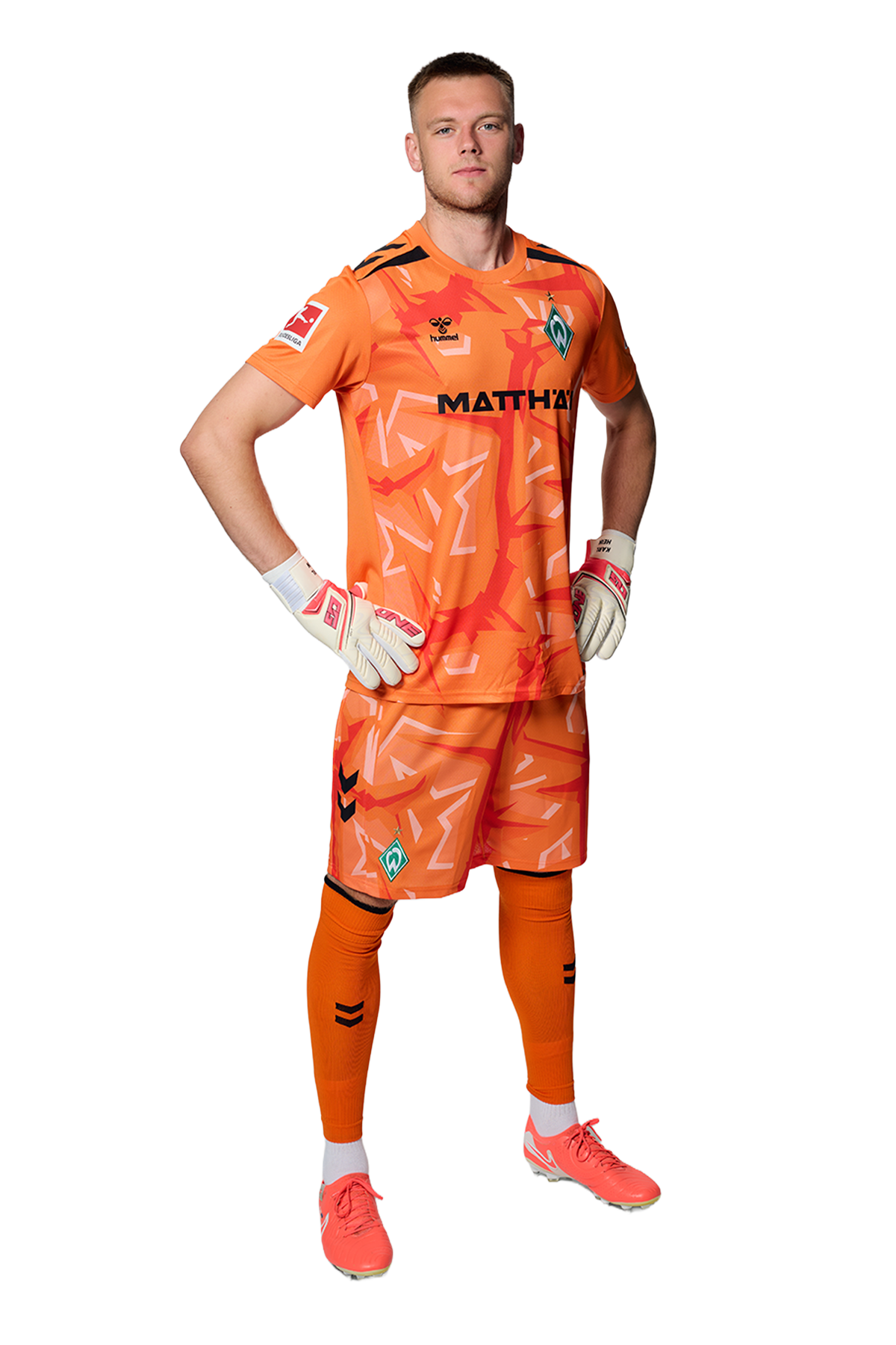 Ein Mann in einem orangenen Fußballtrikot von Werder Bremen steht selbstbewusst mit den Händen in den Hüften vor einem einfarbigen Hintergrund.