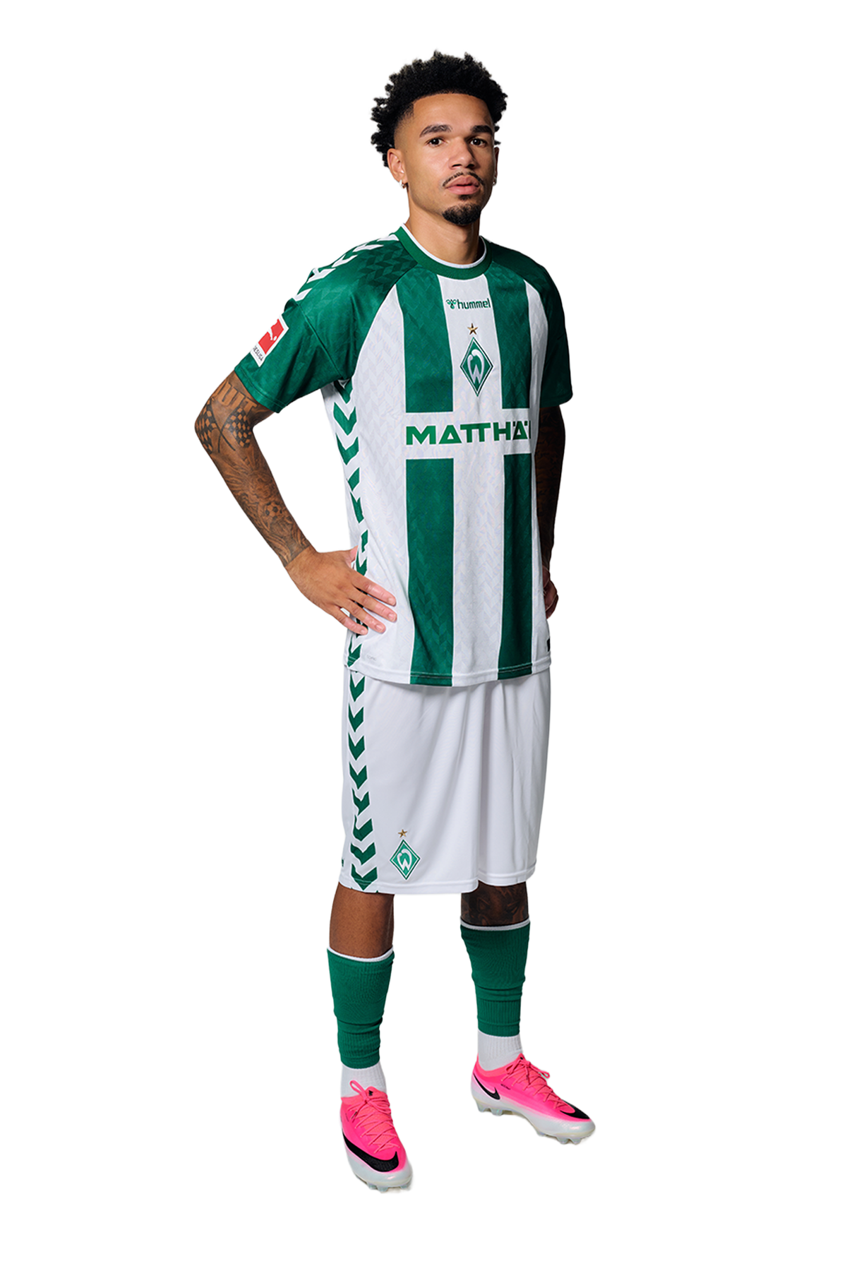 Ein Mann in einem grün-weiß gestreiften Fußballtrikot von Werder Bremen steht selbstbewusst mit den Händen in den Hüften vor einem einfarbigen Hintergrund.