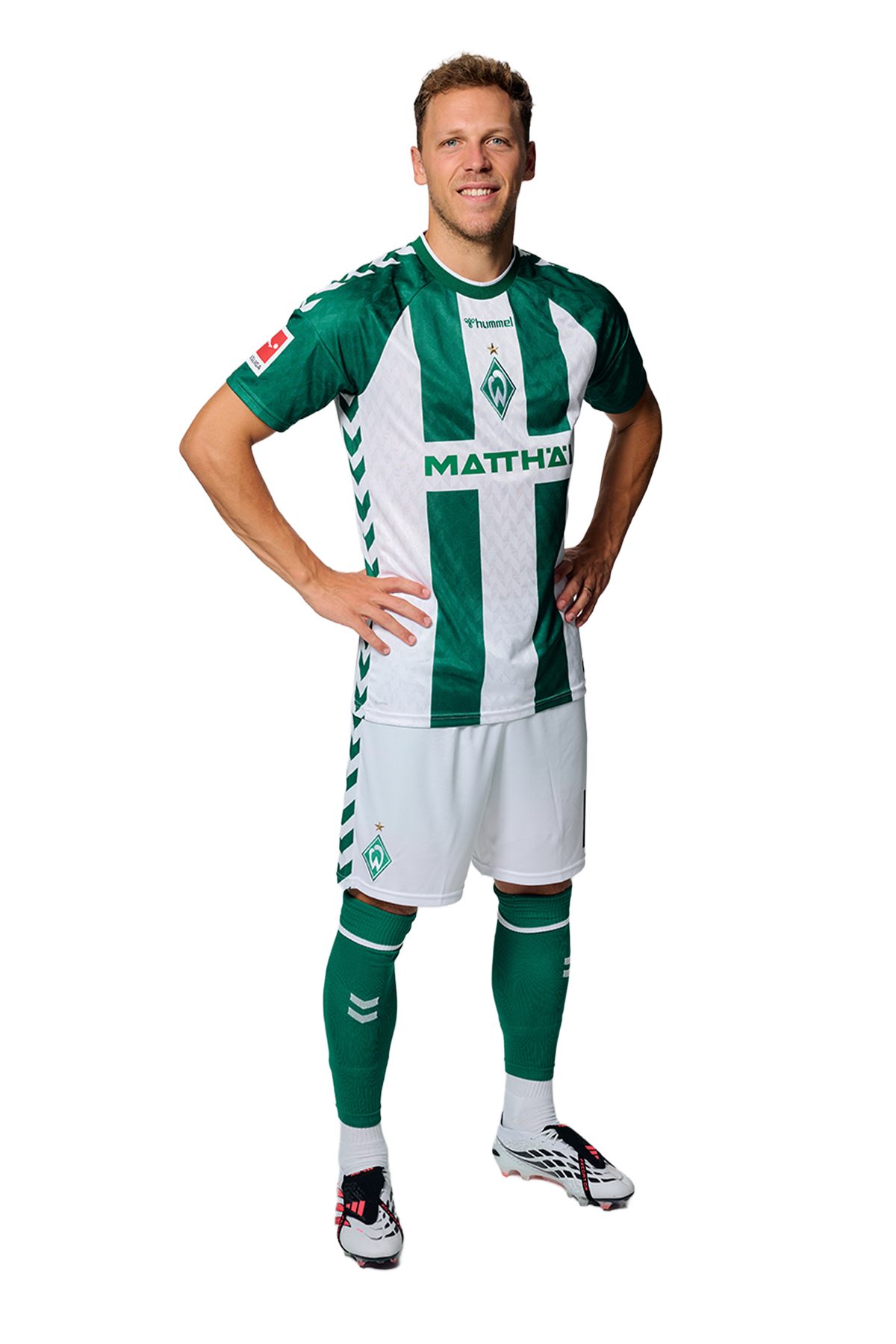 Ein Mann in einem grün-weiß gestreiften Fußballtrikot von Werder Bremen steht selbstbewusst mit den Händen in den Hüften vor einem einfarbigen Hintergrund.