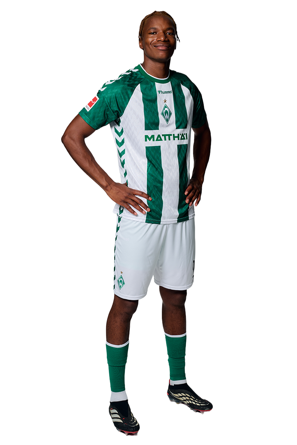 Ein Mann in einem grün-weiß gestreiften Fußballtrikot von Werder Bremen steht selbstbewusst mit den Händen in den Hüften vor einem einfarbigen Hintergrund.