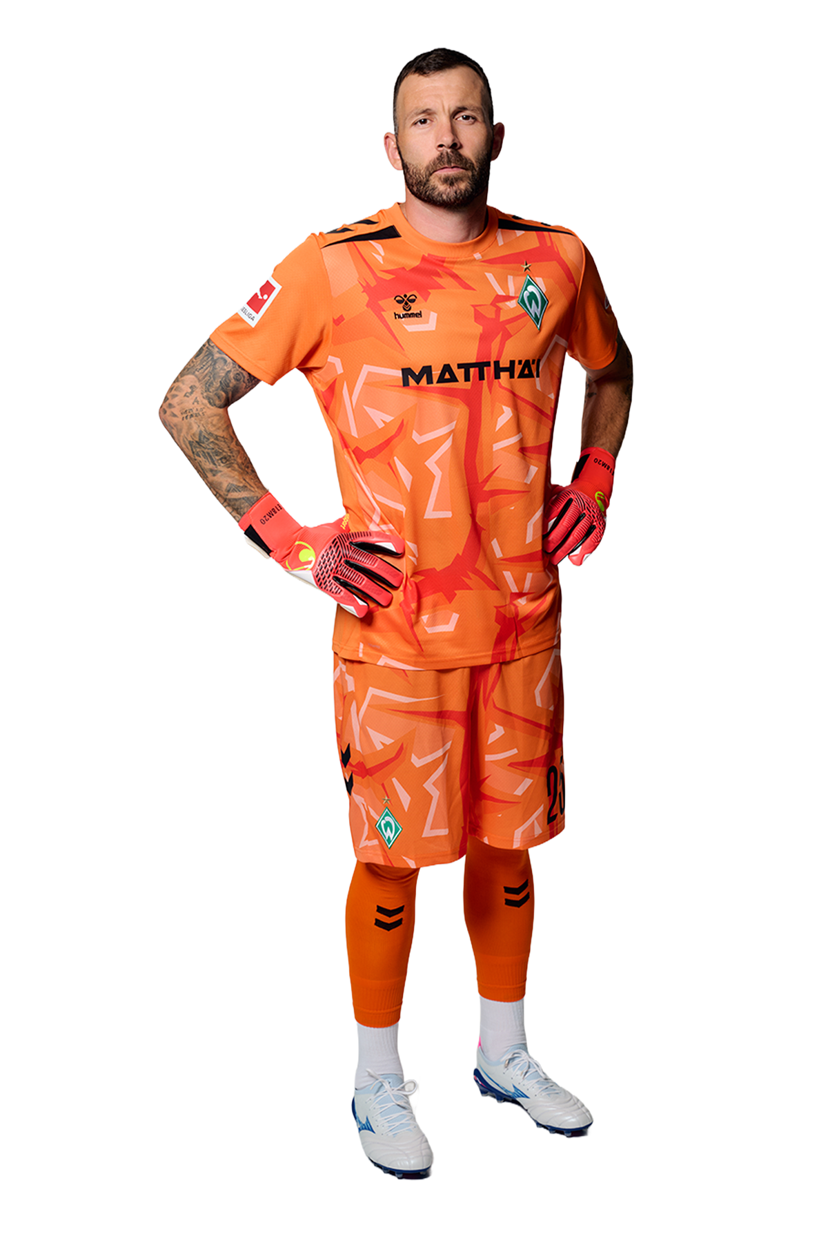Ein Mann in einem orangenen Fußballtrikot von Werder Bremen steht selbstbewusst mit den Händen in den Hüften vor einem einfarbigen Hintergrund.
