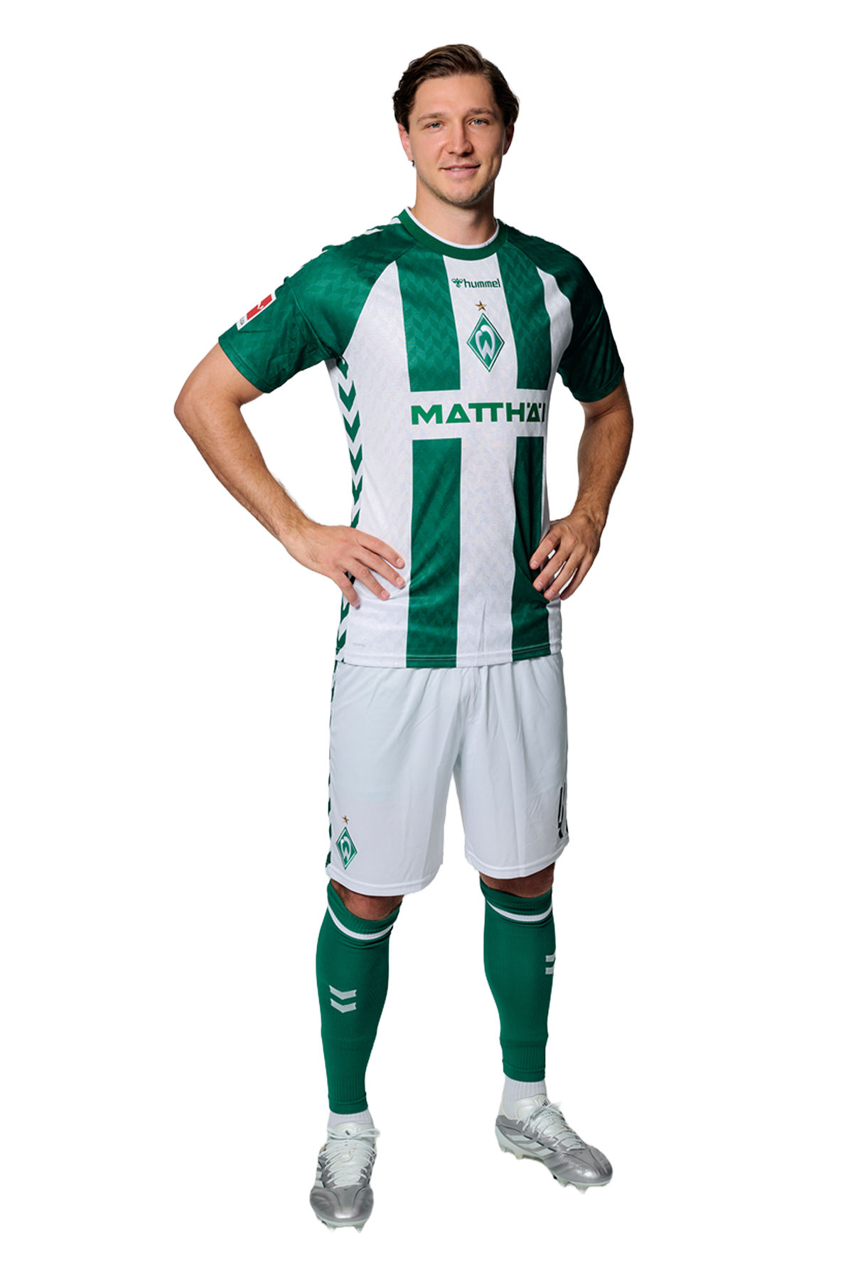 Ein Mann in einem grün-weiß gestreiften Fußballtrikot von Werder Bremen steht selbstbewusst mit den Händen in den Hüften vor einem einfarbigen Hintergrund.