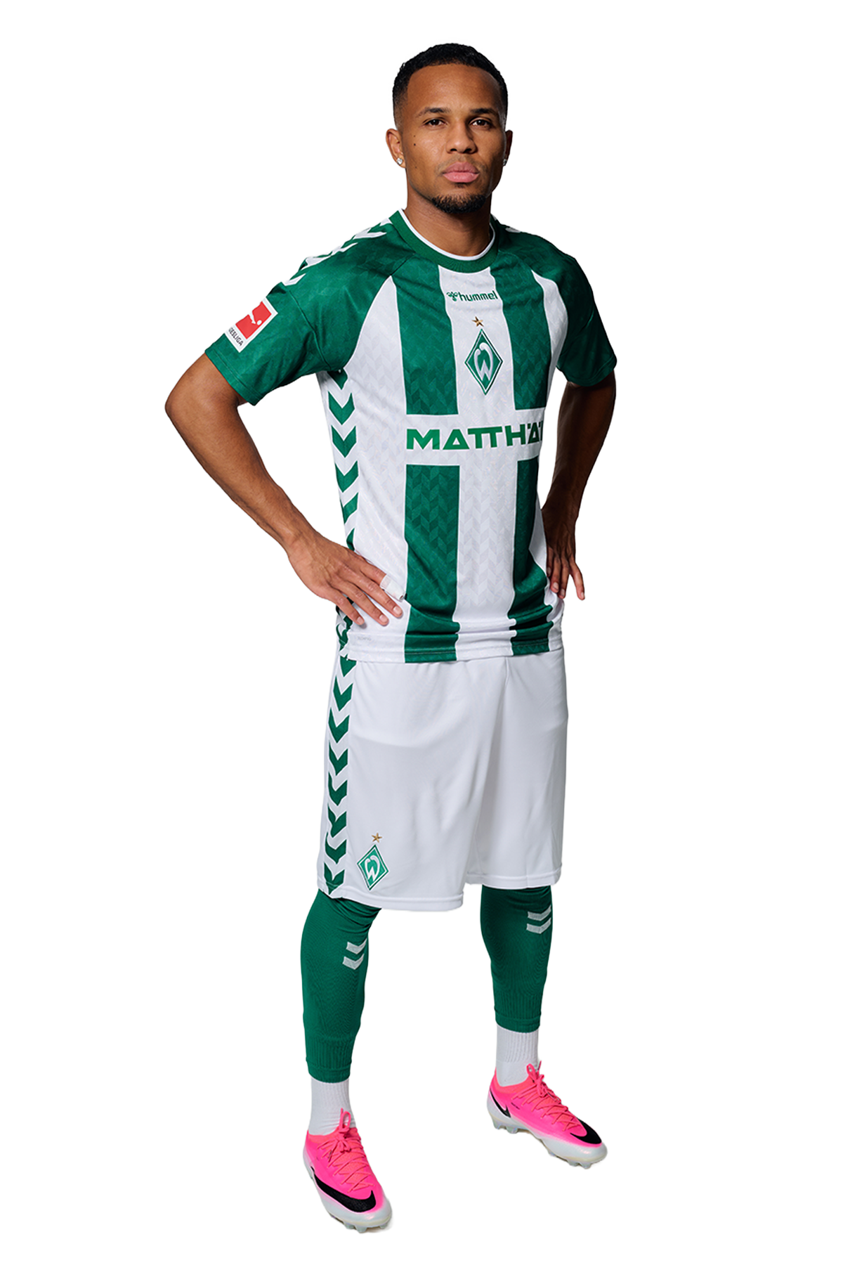 Ein Mann in einem grün-weiß gestreiften Fußballtrikot von Werder Bremen steht selbstbewusst mit den Händen in den Hüften vor einem einfarbigen Hintergrund.