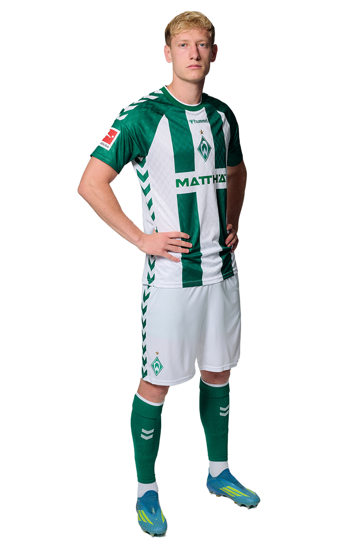Ein Mann in einem grün-weiß gestreiften Fußballtrikot von Werder Bremen steht selbstbewusst mit den Händen in den Hüften vor einem einfarbigen Hintergrund.