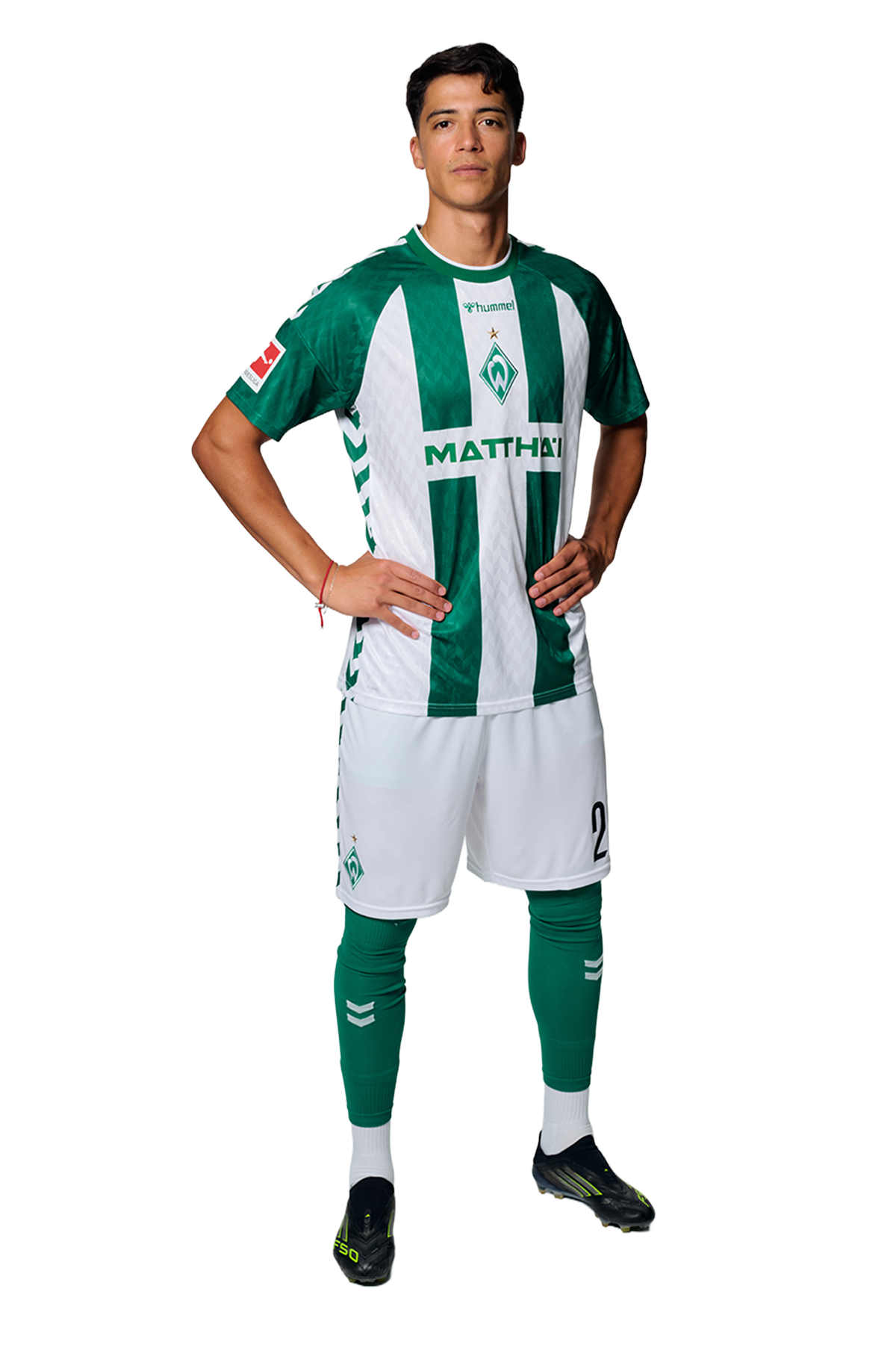 Ein Mann in einem grün-weiß gestreiften Fußballtrikot von Werder Bremen steht selbstbewusst mit den Händen in den Hüften vor einem einfarbigen Hintergrund.