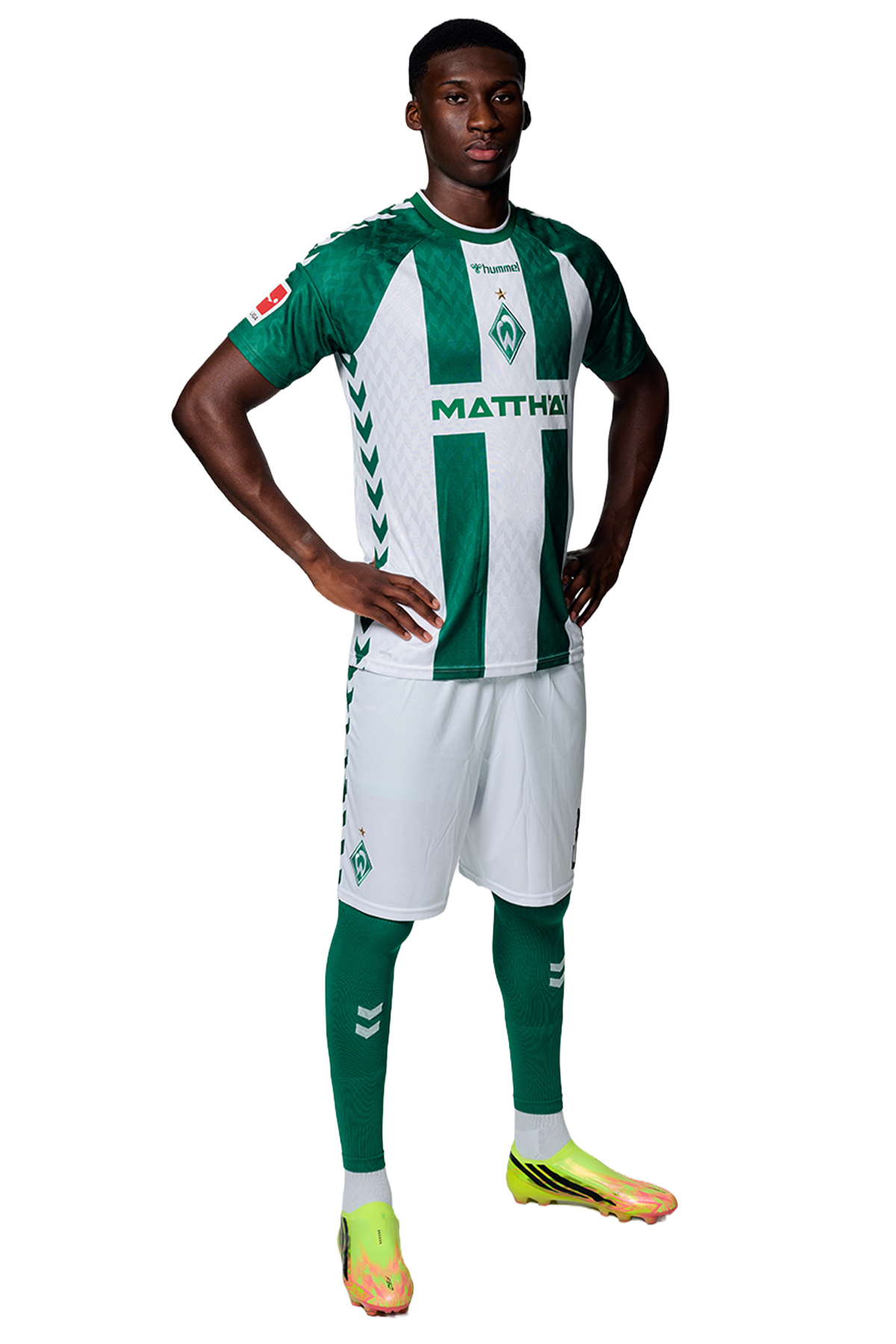 Ein Mann in einem grün-weiß gestreiften Fußballtrikot von Werder Bremen steht selbstbewusst mit den Händen in den Hüften vor einem einfarbigen Hintergrund.