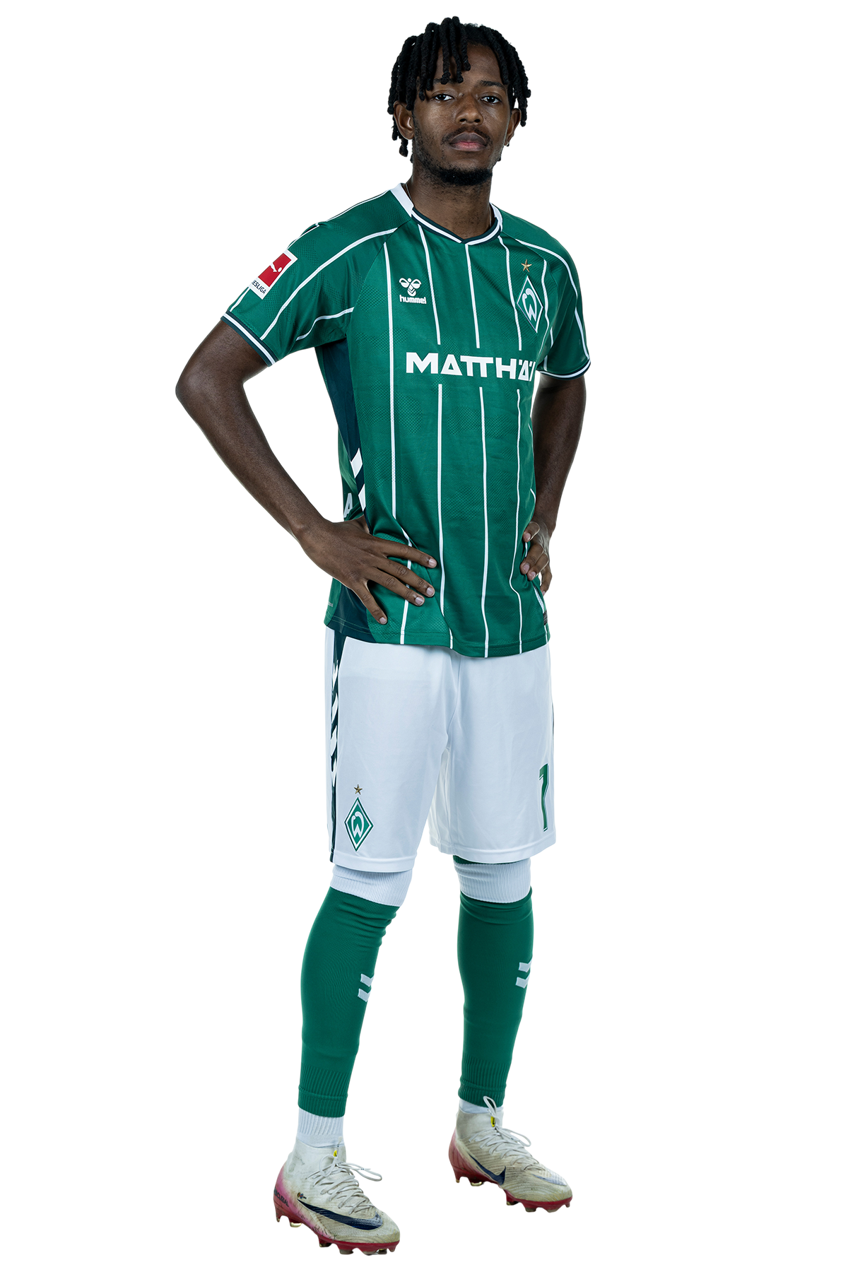 Samuel Mbangula in einem grün-weiß gestreiften Trikot mit der Aufschrift „MATTHÄI“ auf der Vorderseite steht mit den Händen in den Hüften da und schaut nach vorne.