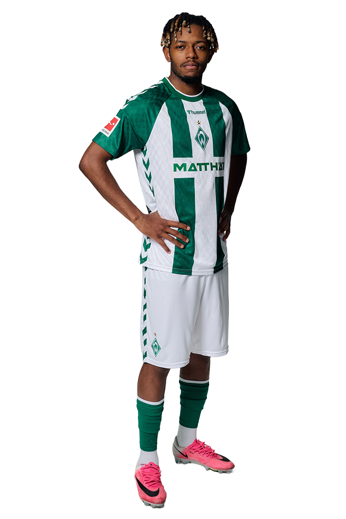 Ein Mann in einem grün-weiß gestreiften Fußballtrikot von Werder Bremen steht selbstbewusst mit den Händen in den Hüften vor einem einfarbigen Hintergrund.