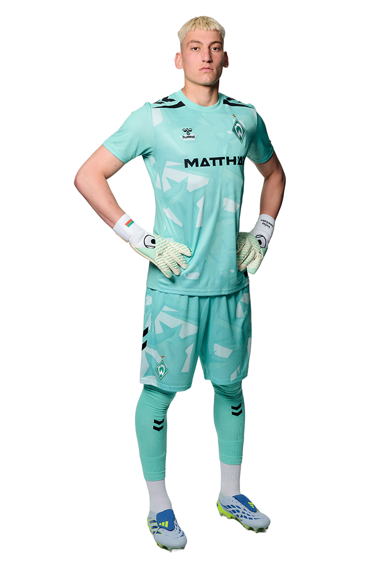 Ein Mann in einem blauen Fußballtrikot von Werder Bremen steht selbstbewusst mit den Händen in den Hüften vor einem einfarbigen Hintergrund.