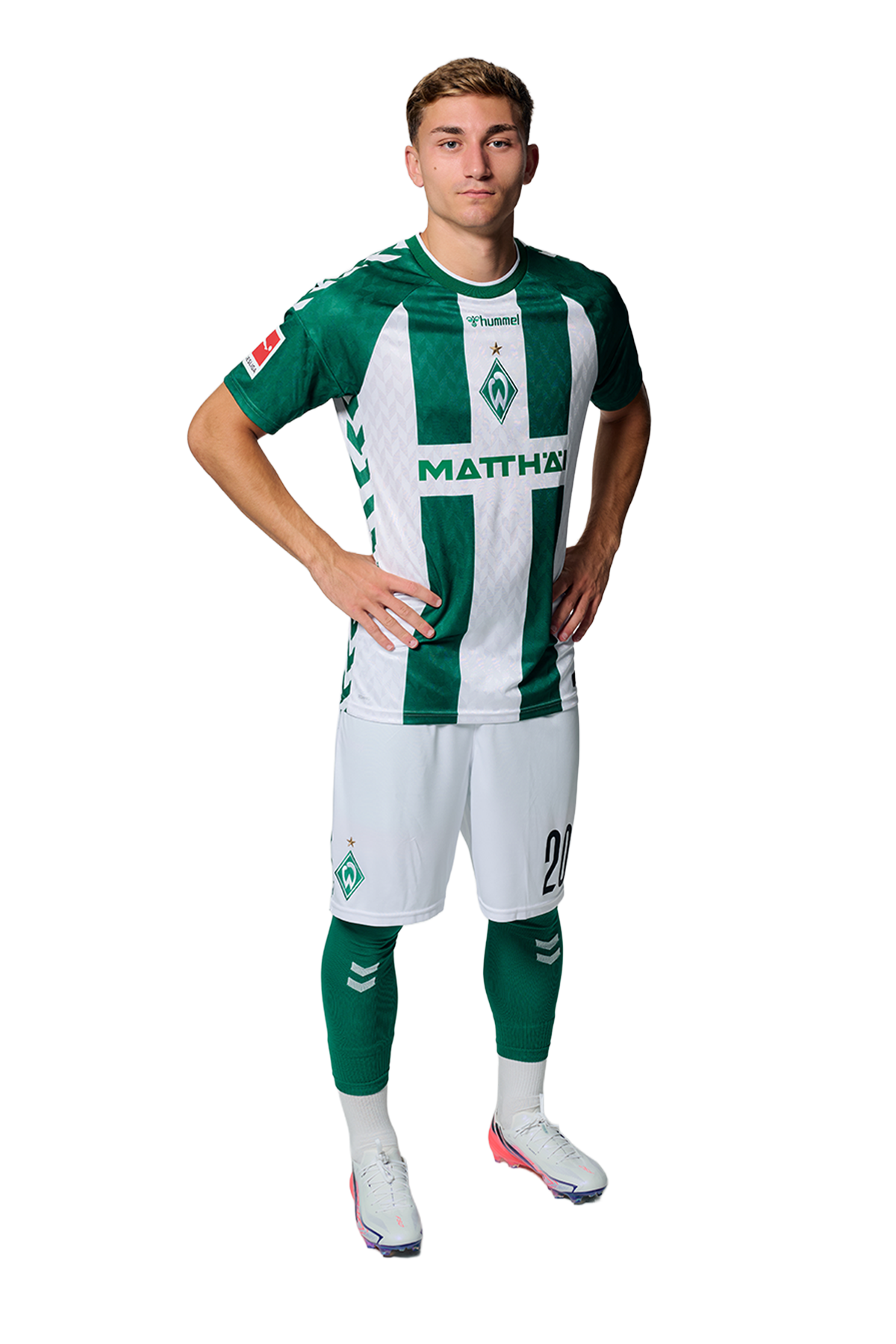Ein Mann in einem grün-weiß gestreiften Fußballtrikot von Werder Bremen steht selbstbewusst mit den Händen in den Hüften vor einem einfarbigen Hintergrund.