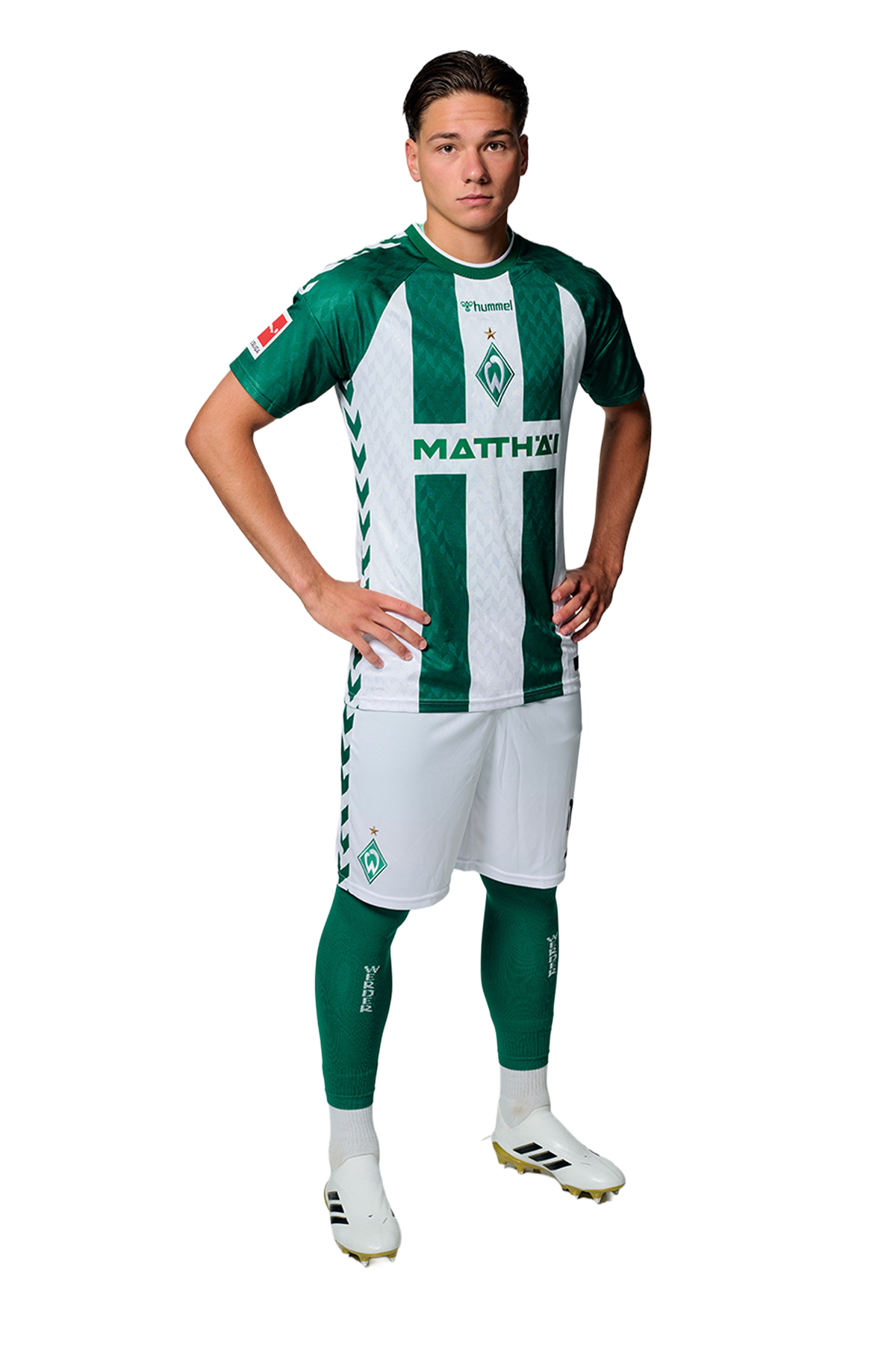 Ein Mann in einem grün-weiß gestreiften Fußballtrikot von Werder Bremen steht selbstbewusst mit den Händen in den Hüften vor einem einfarbigen Hintergrund.