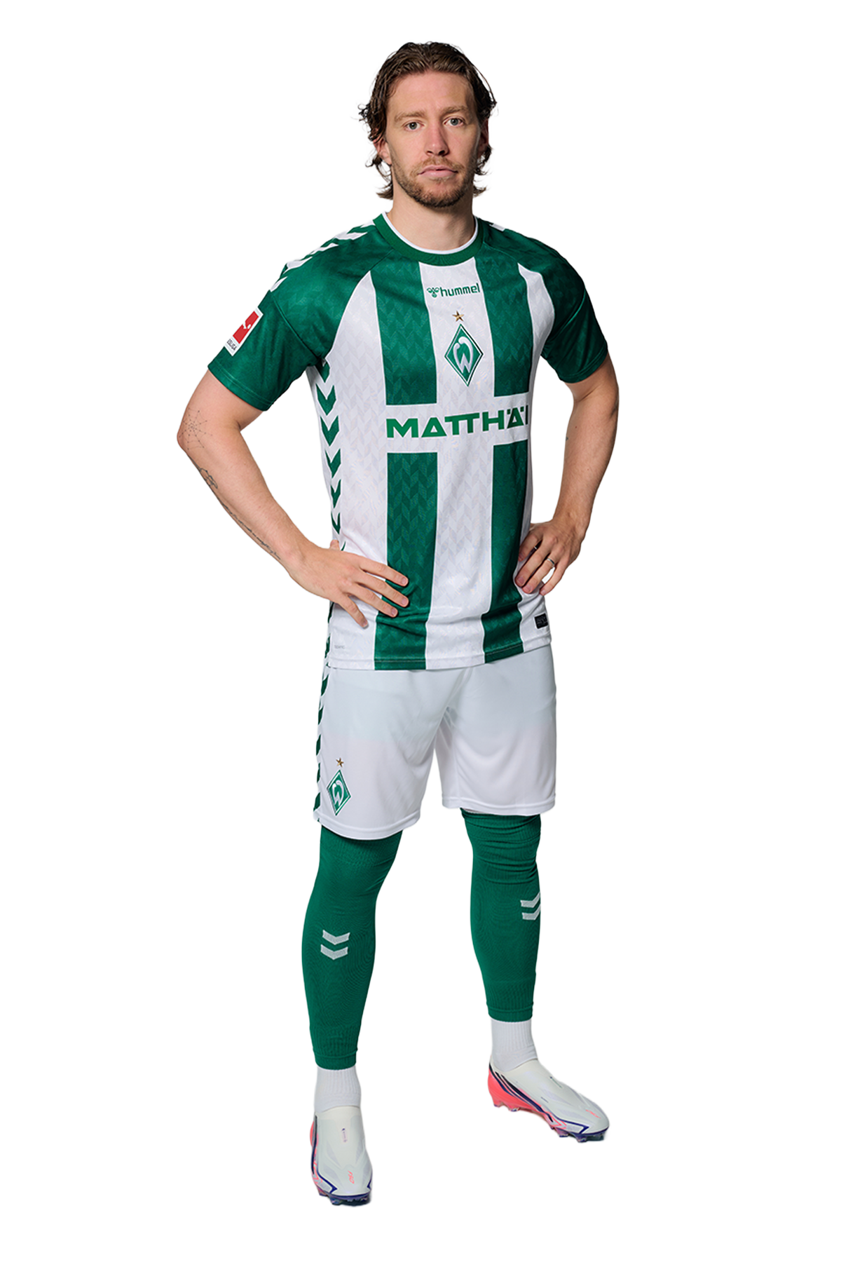 Ein Mann in einem grün-weiß gestreiften Fußballtrikot von Werder Bremen steht selbstbewusst mit den Händen in den Hüften vor einem einfarbigen Hintergrund.