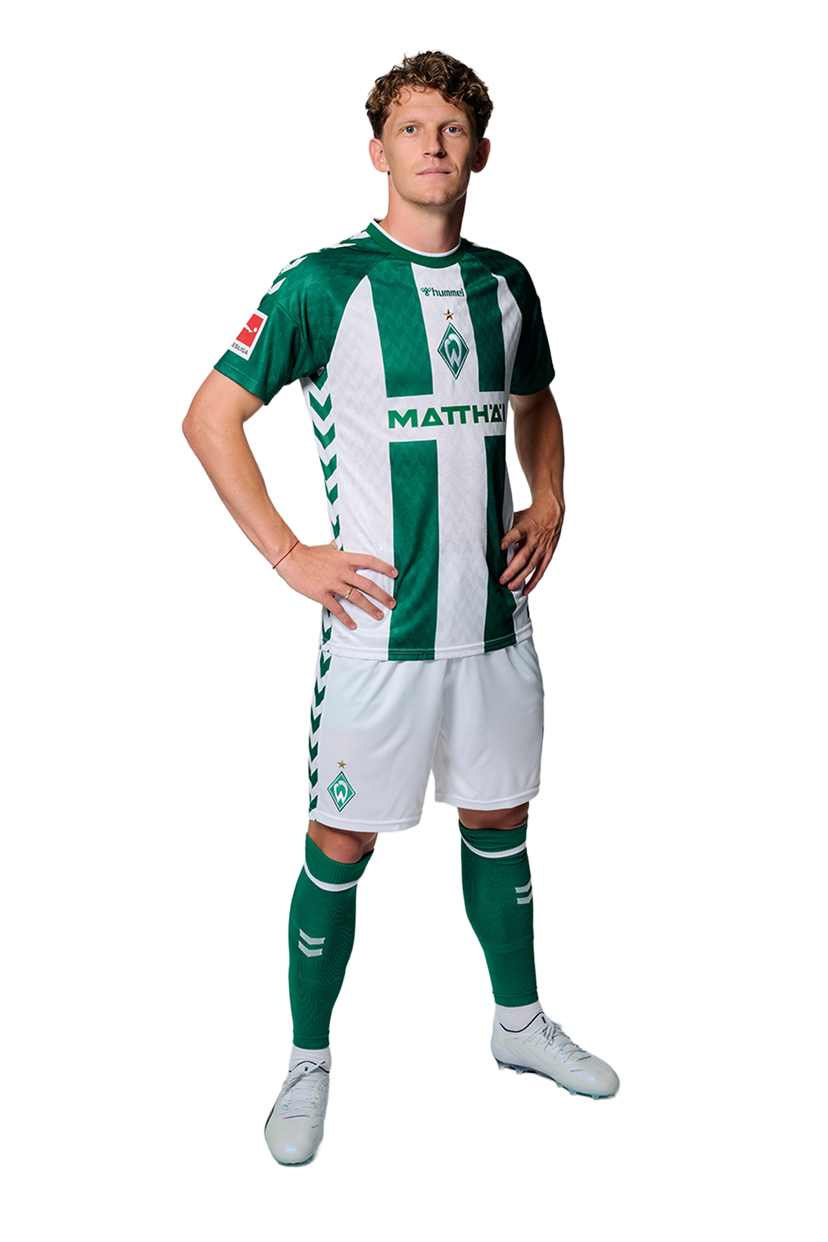 Ein Mann in einem grün-weiß gestreiften Fußballtrikot von Werder Bremen steht selbstbewusst mit den Händen in den Hüften vor einem einfarbigen Hintergrund.