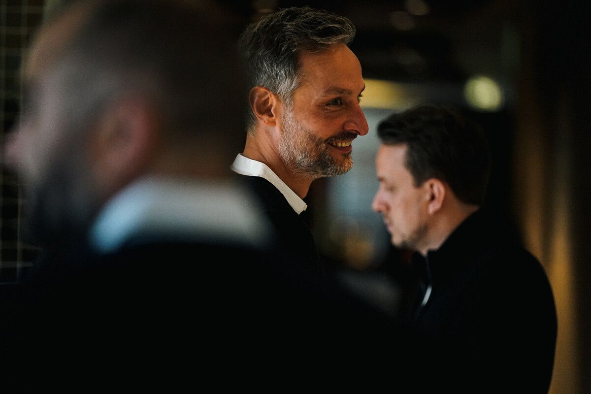 Drei Männer in formeller Kleidung stehen in einem schwach beleuchteten Raum, wobei ein Mann im Fokus steht, lächelt und zur Seite schaut.