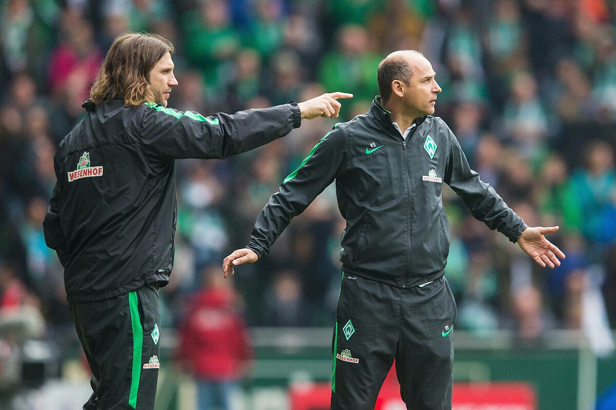 Zwei Trainer in schwarzen Sportjacken auf einem Fußballfeld, einer zeigt mit dem Finger, der andere hat die Arme ausgestreckt, im Hintergrund eine verschwommene Menschenmenge.