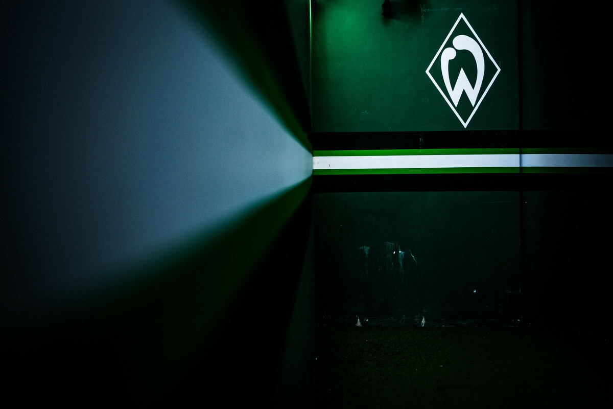 Dunkler Flur mit grüner Beleuchtung, an der Wand ein weißes geometrisches Logo. Reflektierender Boden und diagonale Lichtstreifen.