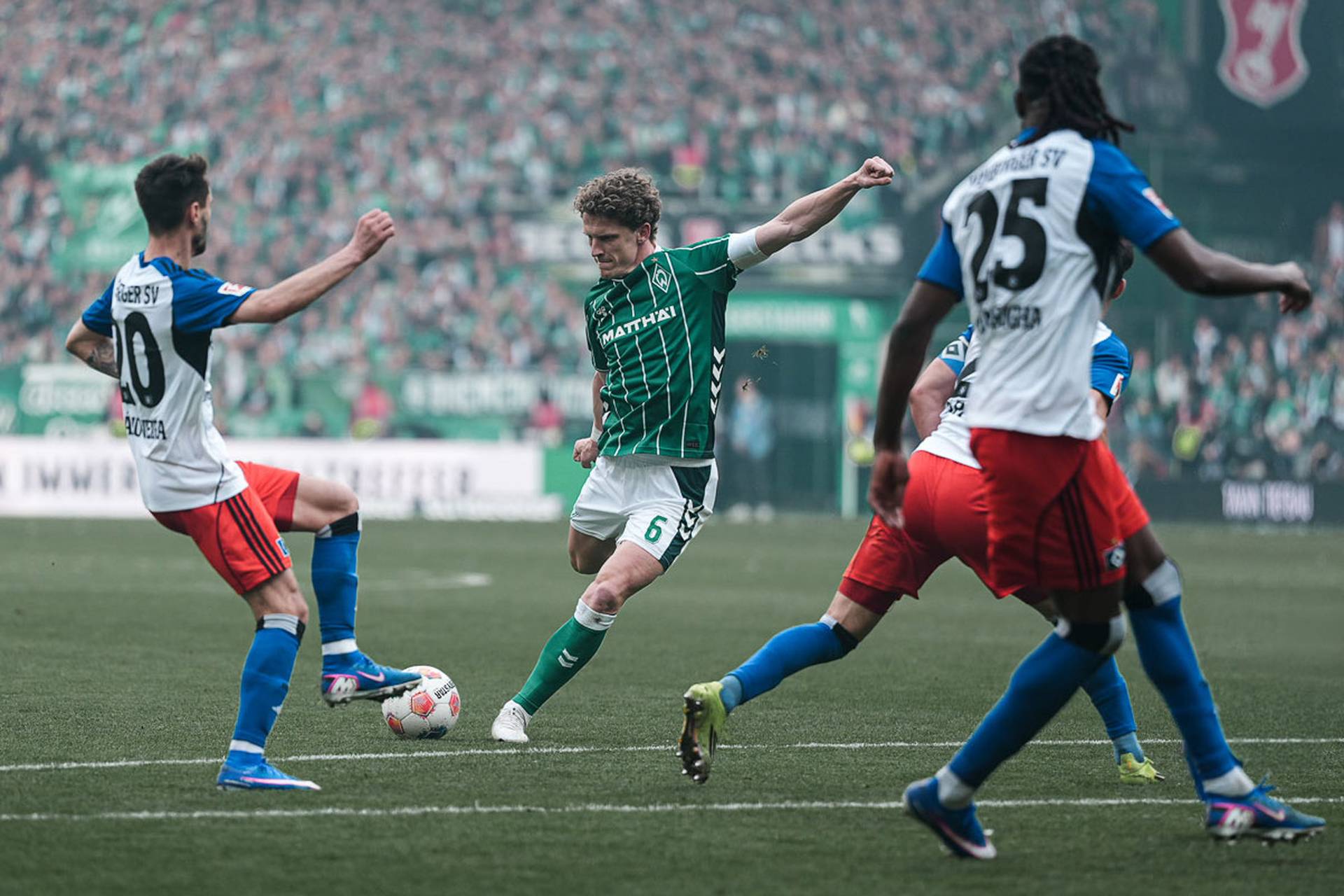 Ein Fußballspieler im grünen Trikot schießt während eines spannenden Spiels auf das Tor, umringt von drei Gegnern in weißen und roten Trikots.