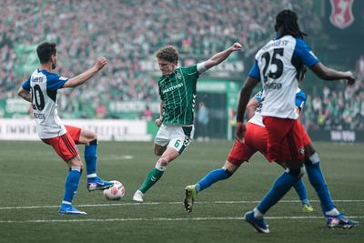 Ein Fußballspieler im grünen Trikot schießt während eines spannenden Spiels auf das Tor, umringt von drei Gegnern in weißen und roten Trikots.