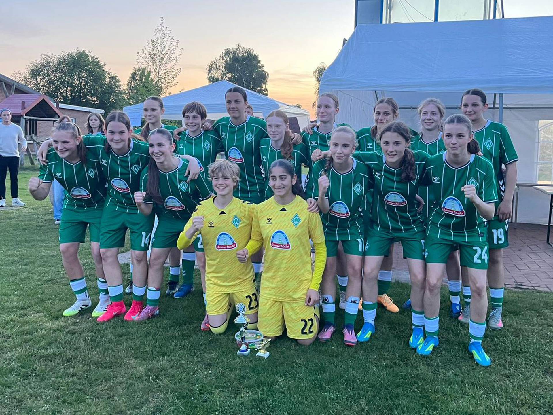 Eine Mädchenfußballmannschaft in grünen Trikots posiert gemeinsam im Freien, wobei zwei Spielerinnen vorne einen Pokal in den Händen halten.