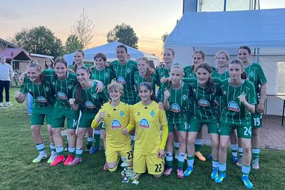 Eine Mädchenfußballmannschaft in grünen Trikots posiert gemeinsam im Freien, wobei zwei Spielerinnen vorne einen Pokal in den Händen halten.