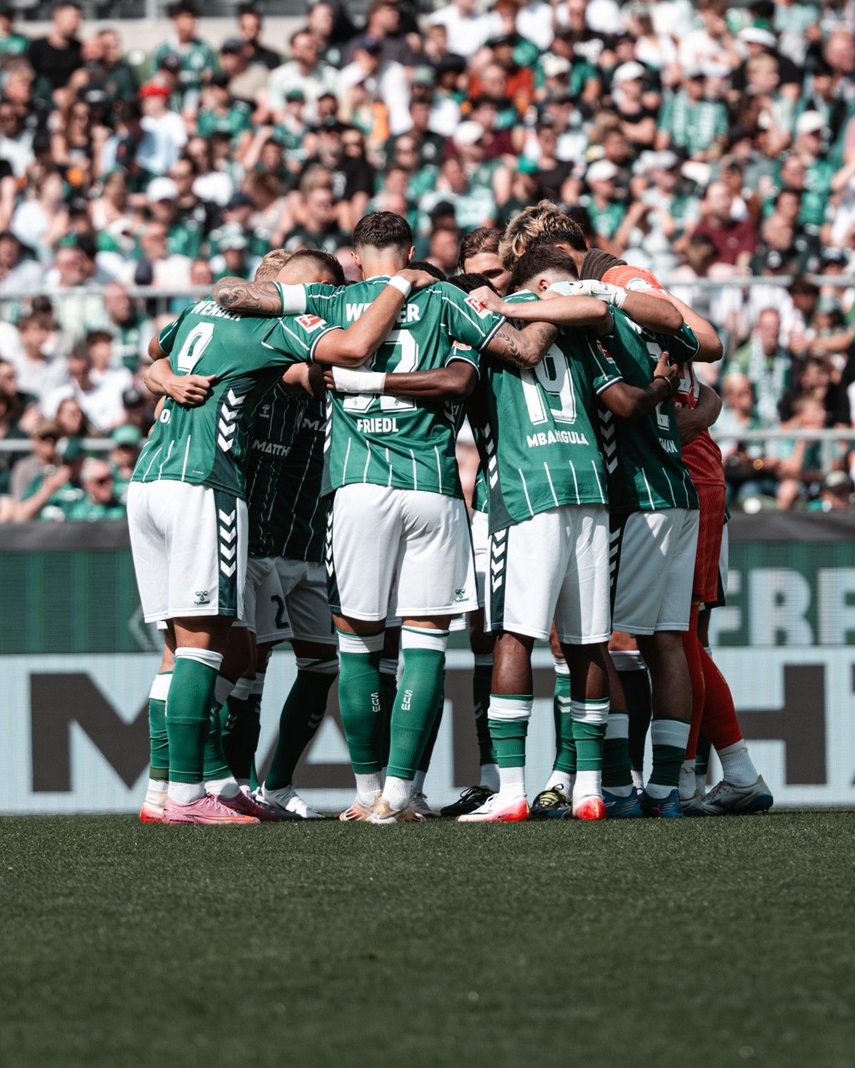 Die Fußballmannschaft in grünen Trikots versammelt sich auf dem Spielfeld, umgeben von einem vollbesetzten Stadion mit Zuschauern.