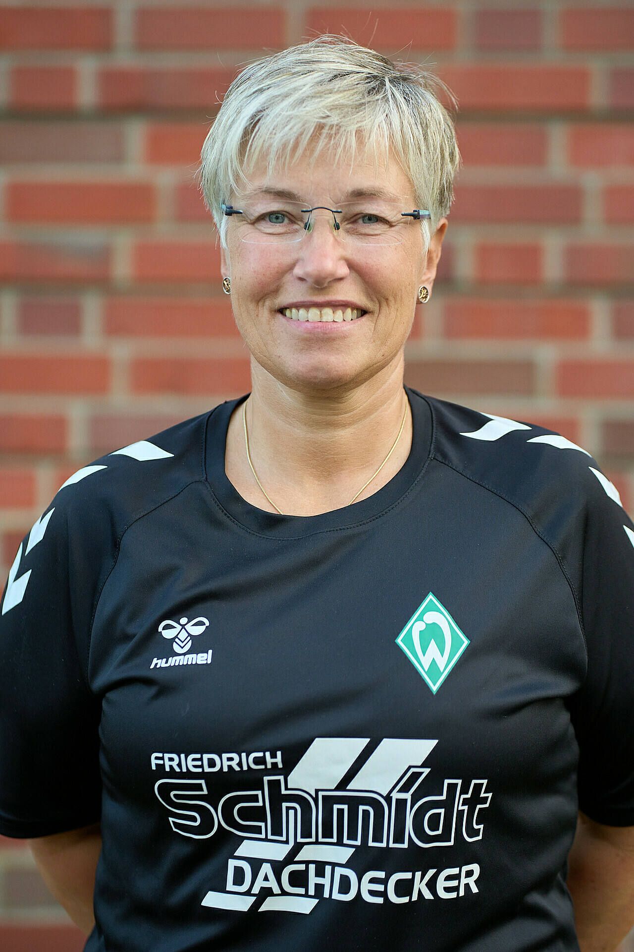 Lächelnde Frau im schwarzem Sportshirt mit Logos, die vor einer Backsteinmauer steht.