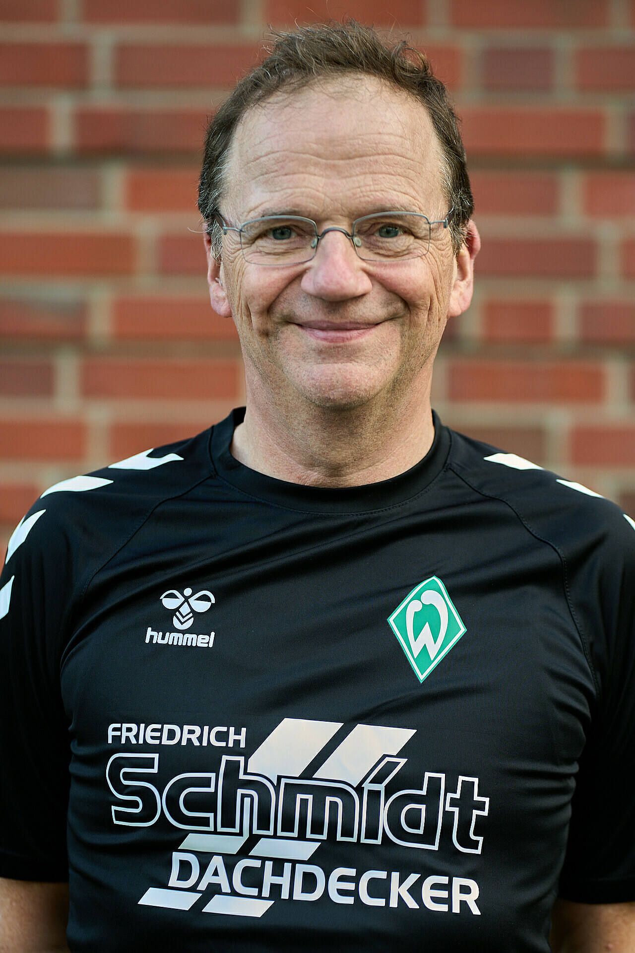 Lächelnder Mann mit Brille und schwarzem Sportshirt mit Logos, der vor einer Backsteinmauer steht.