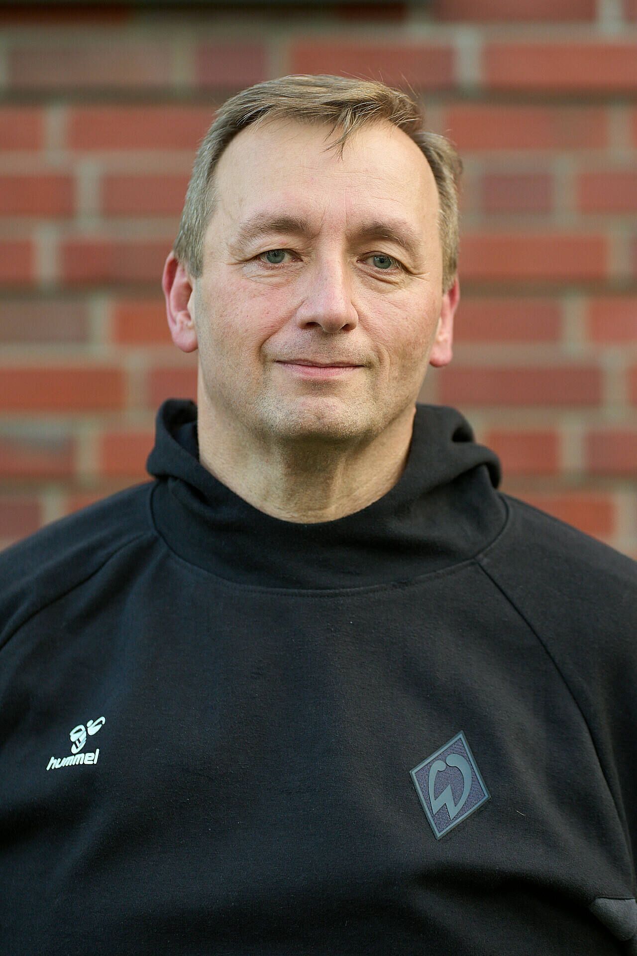Lächelnder Mann im schwarzem Hoodie mit Logos, der vor einer Backsteinmauer steht.