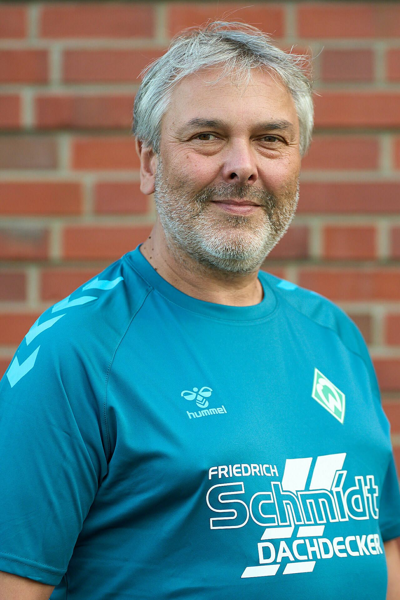 Lächelnder Mann im blauem Sportshirt mit Logos, der vor einer Backsteinmauer steht.