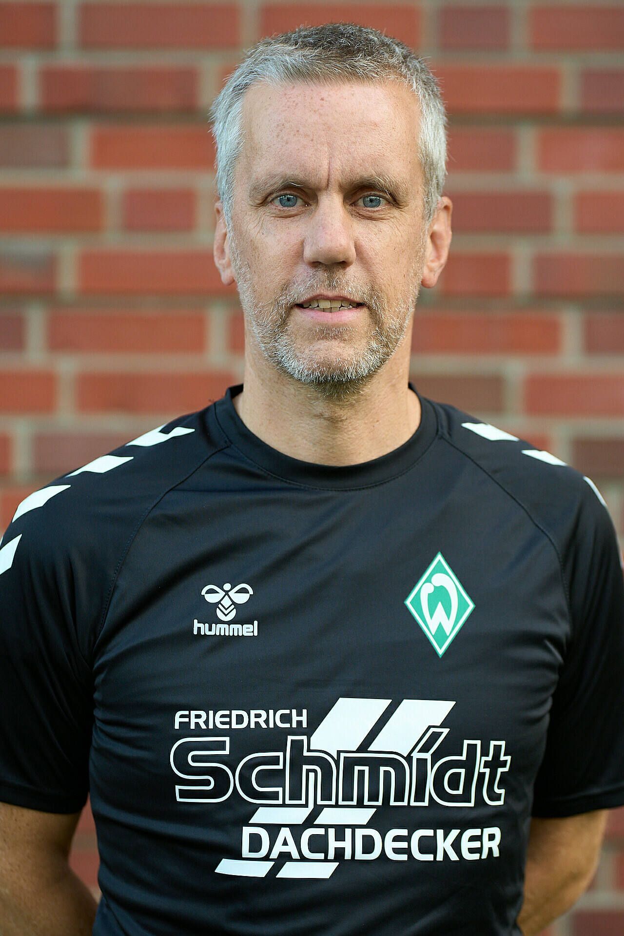 Lächelnder Mann im schwarzem Sportshirt mit Logos, der vor einer Backsteinmauer steht.